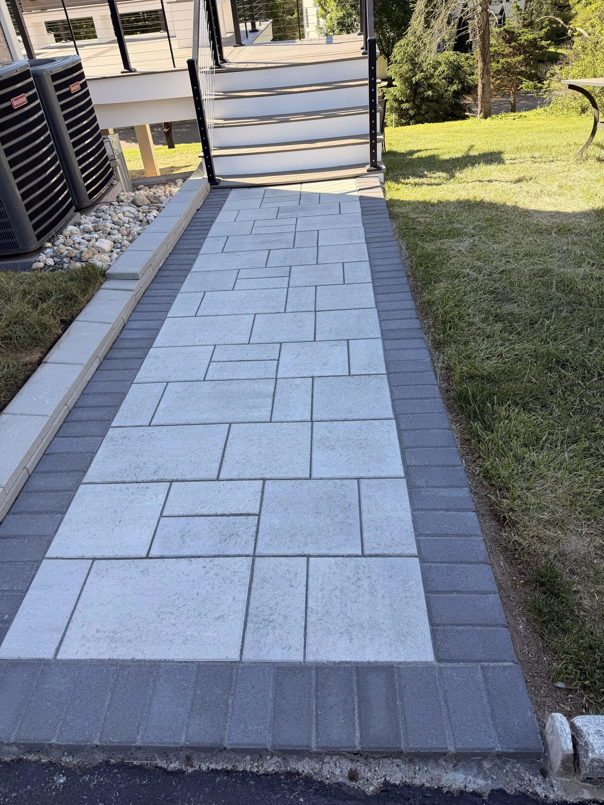 A paved walkway with large rectangular light gray tiles bordered by dark gray bricks, leading to a set of stairs with a black handrail, next to a grassy yard with trees and a wooden deck in the background.