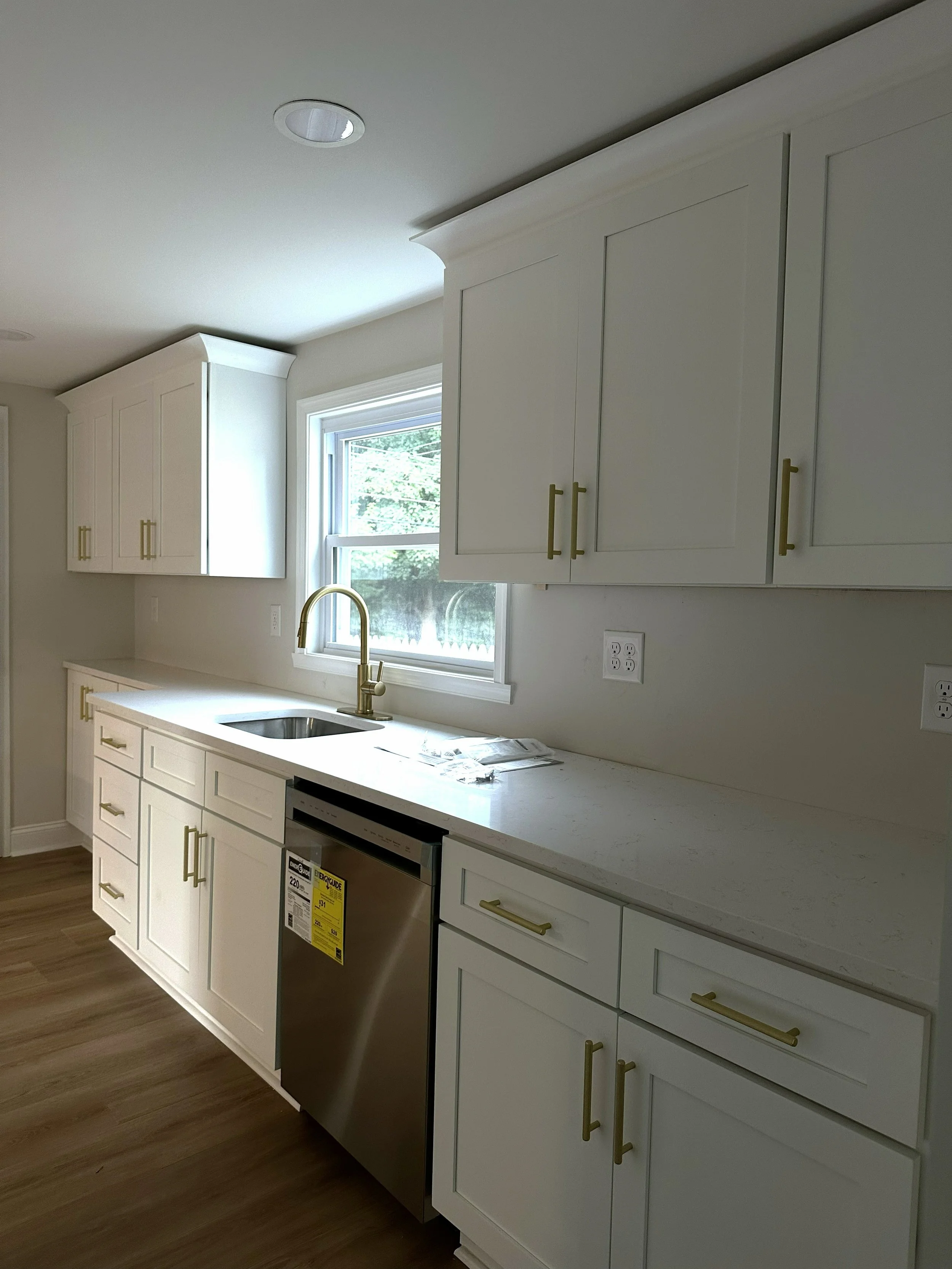 Modern kitchen with white cabinets, gold handles, granite countertop, stainless steel dishwasher, window above the sink, and LVP flooring.