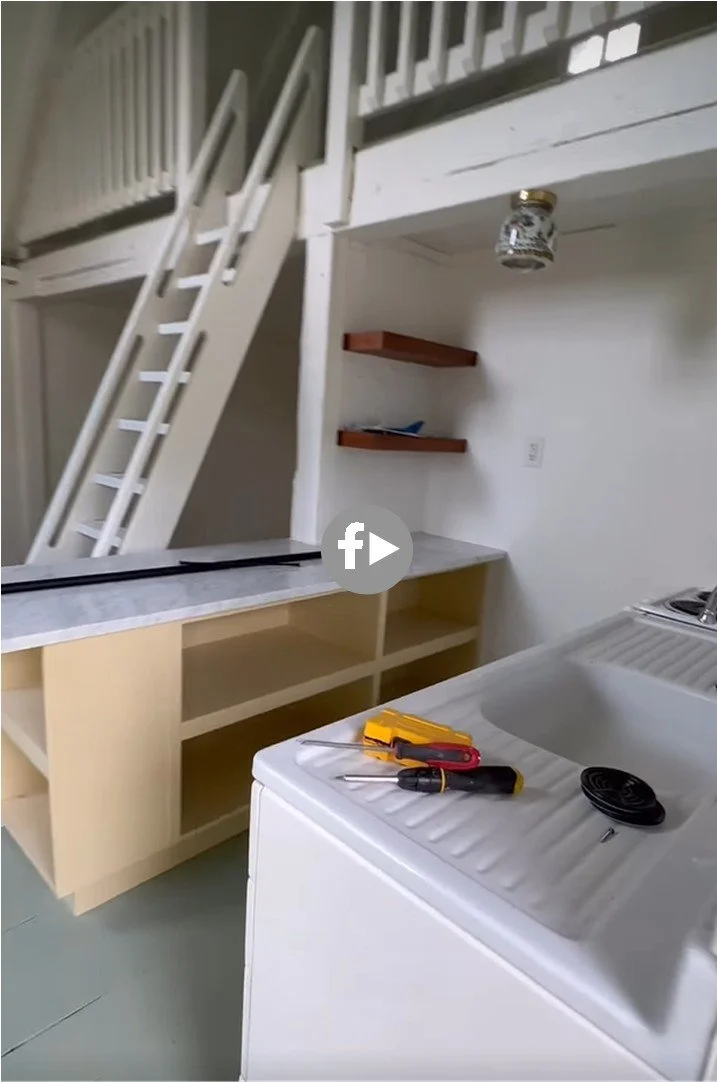 Interior of a small kitchen area with a washing machine, two screwdrivers, a drill, and a utility knife on top. There are open shelves and a countertop, with a loft space above accessible by a white ladder.
