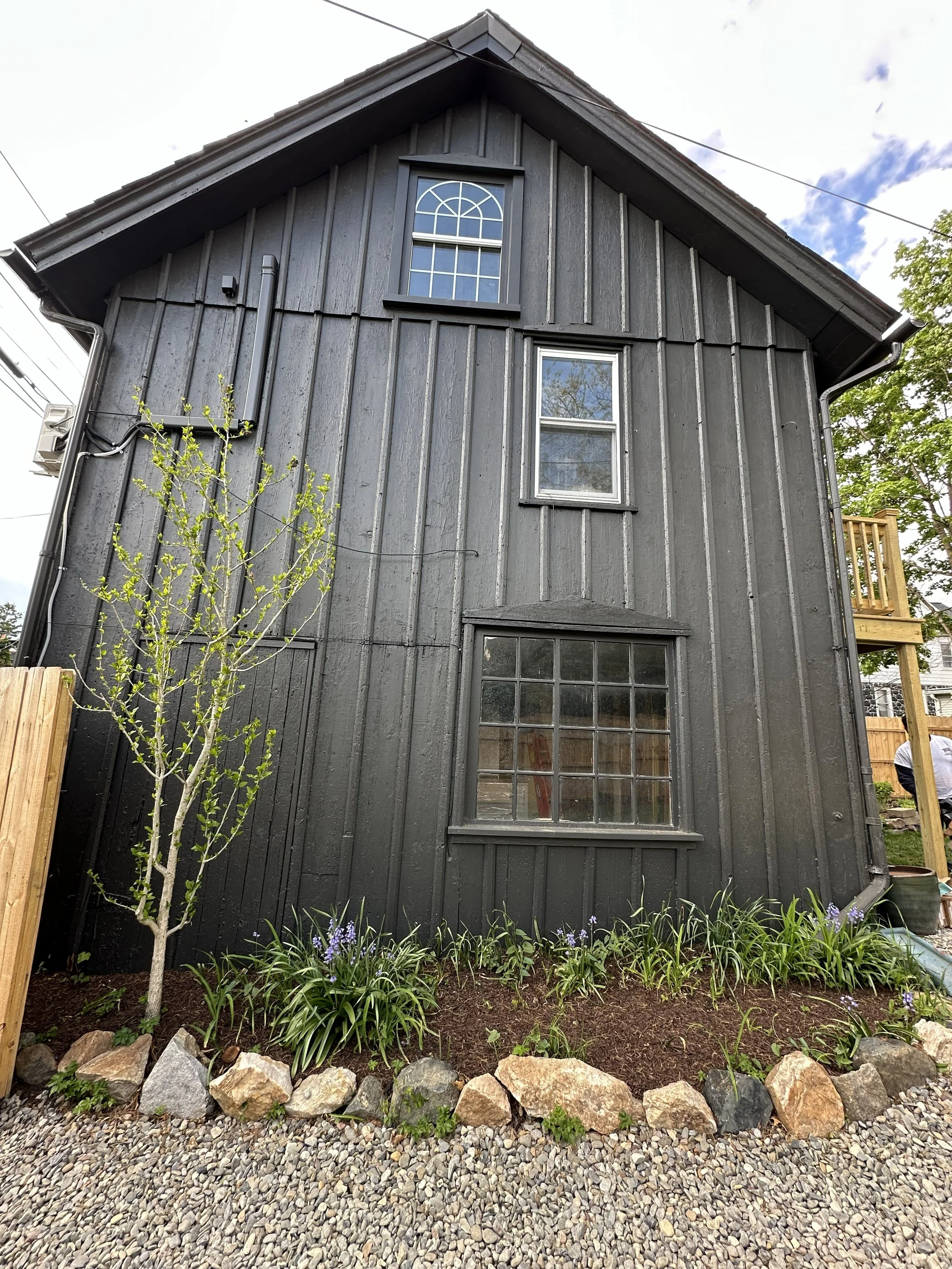 A black wooden house with two windows, one on each floor, and a small garden in front with plants, purple flowers, and rocks, and a gravel pathway.
