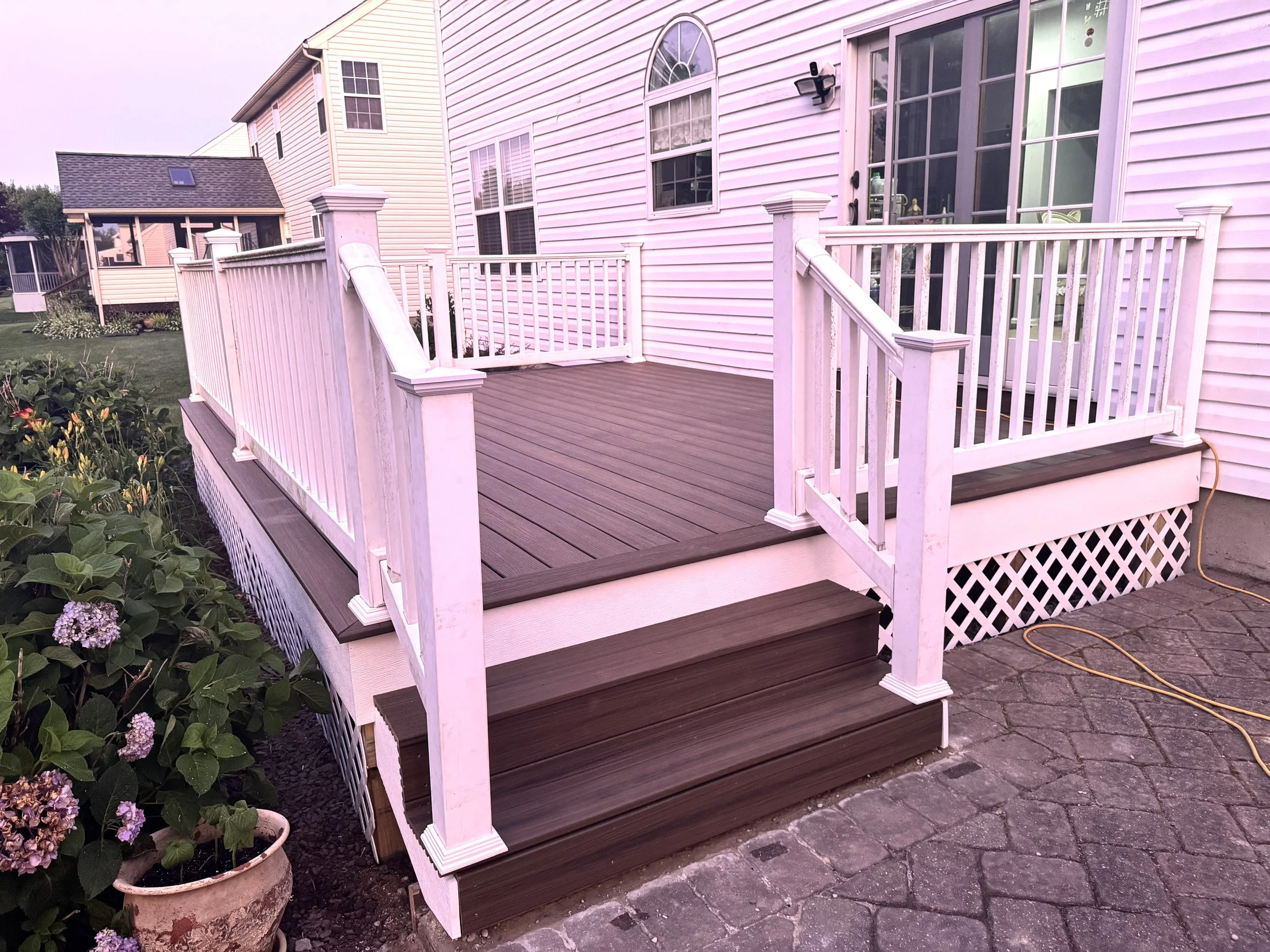 A wooden deck with white railing attached to a house with white siding and large windows, with plants in a flowerpot and a flower bed nearby.