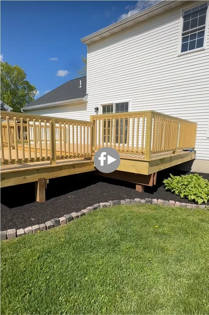 Newly built wooden deck attached to the back of a white house, with fresh black mulch and a small curved edging made of bricks. There is a green shrub and a well-maintained lawn in the yard.