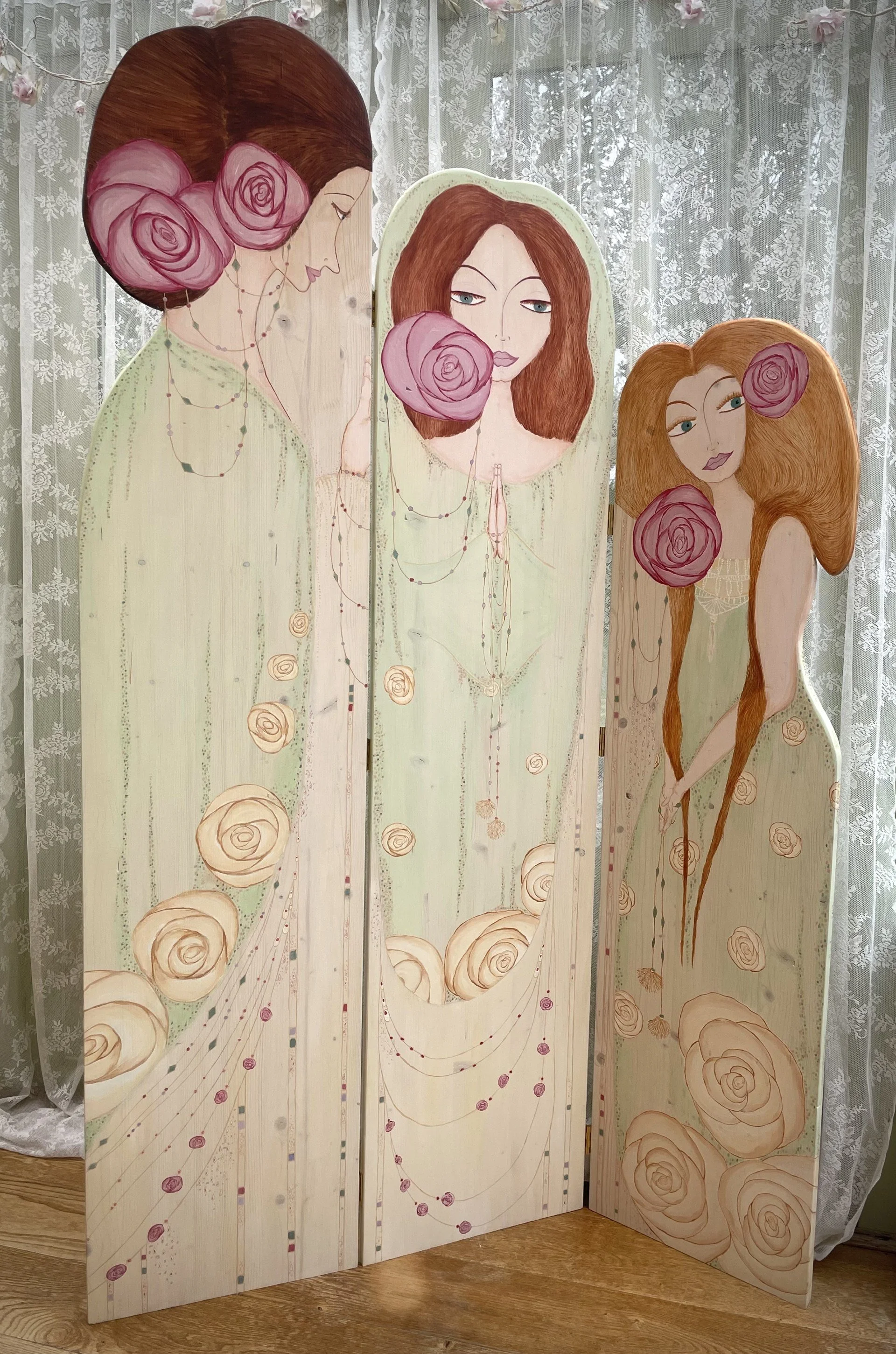 A decorative folding screen featuring artistic portraits of three women with curly brown hair and pink roses. The women have different expressions and hairstyles, with the leftmost woman in profile, the middle woman with hands in prayer, and the rightmost woman with hands crossed. The artwork is painted on a wooden surface with a light background and floral patterns.