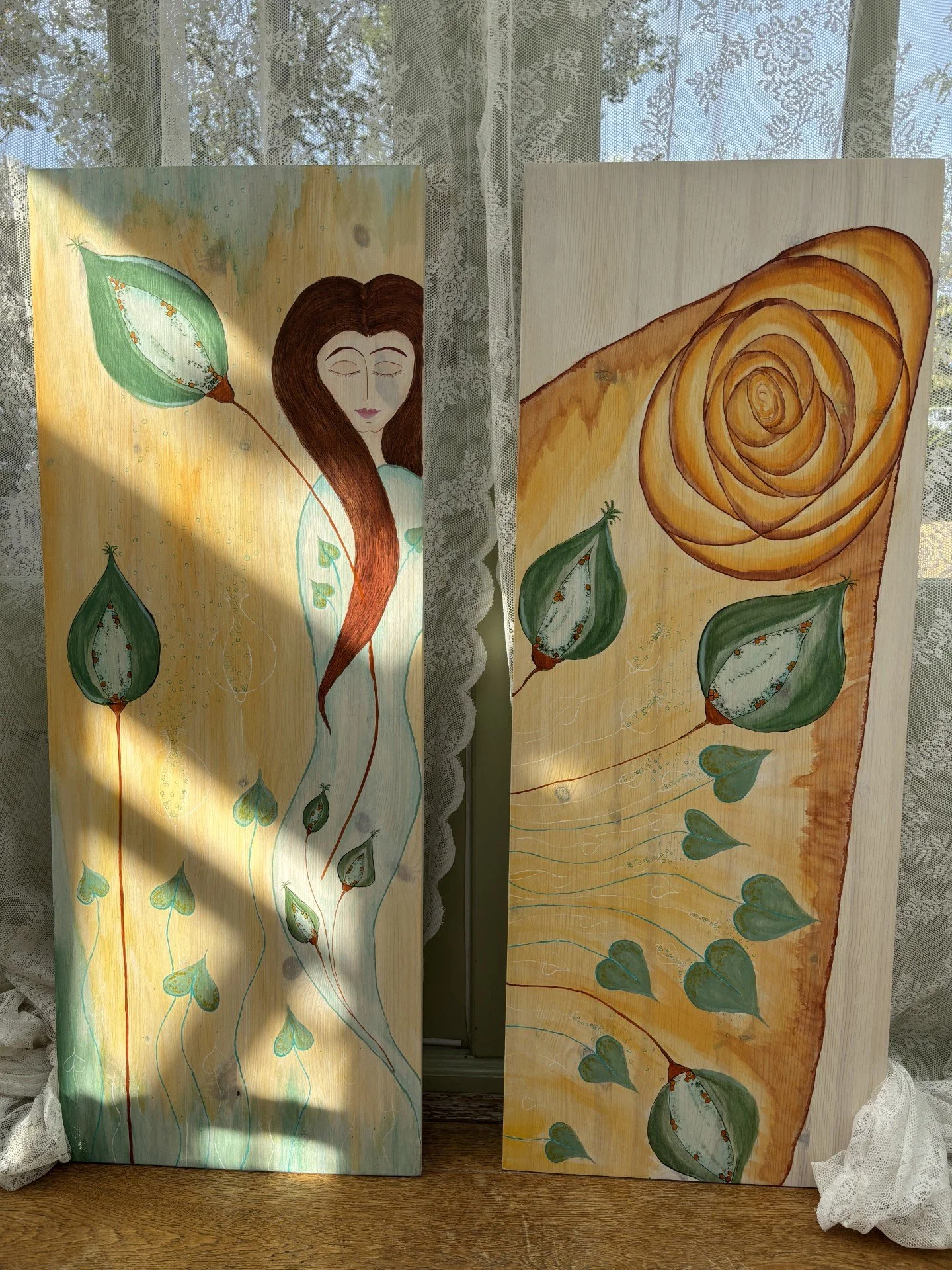 Two painted wooden panels with abstract and figurative artwork, featuring a woman with long brown hair, closed eyes, and green leaves and heart-shaped elements on a yellow background, placed in front of a lace curtain with sunlight shining through.