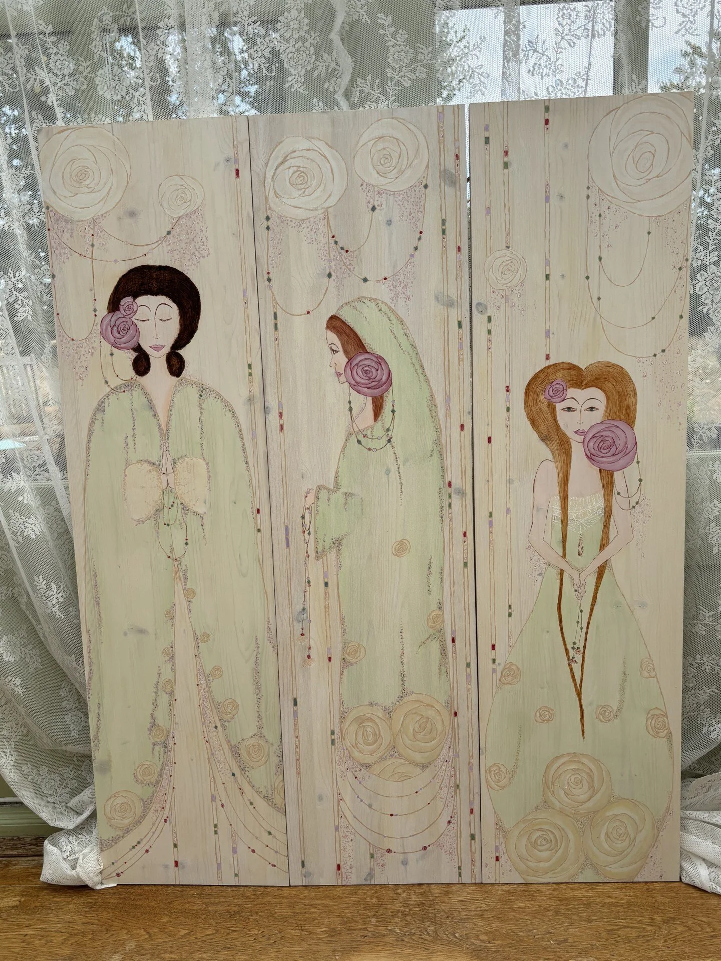 A three-panel wooden artwork featuring three women, each with a large rose near their face and long hair, set against a backdrop of painted roses and hanging decorative elements.