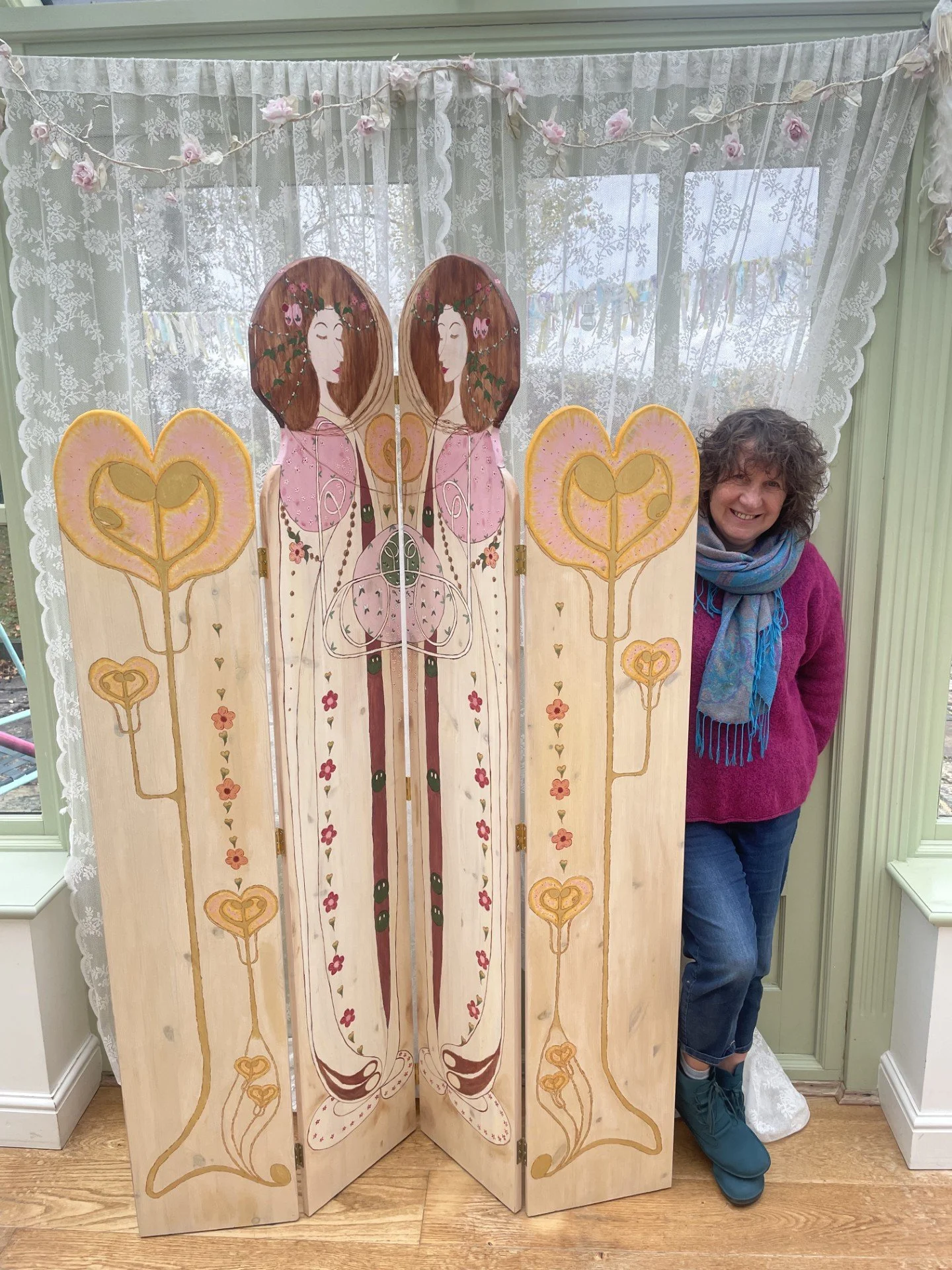 A decorative folding screen, consisting of four panels, featuring mirrored images of Glasgow Style ladies with long brown hair, floral elements, and heart shapes painted on the screen, next to the ladies are warm glowing lantern shaped images.