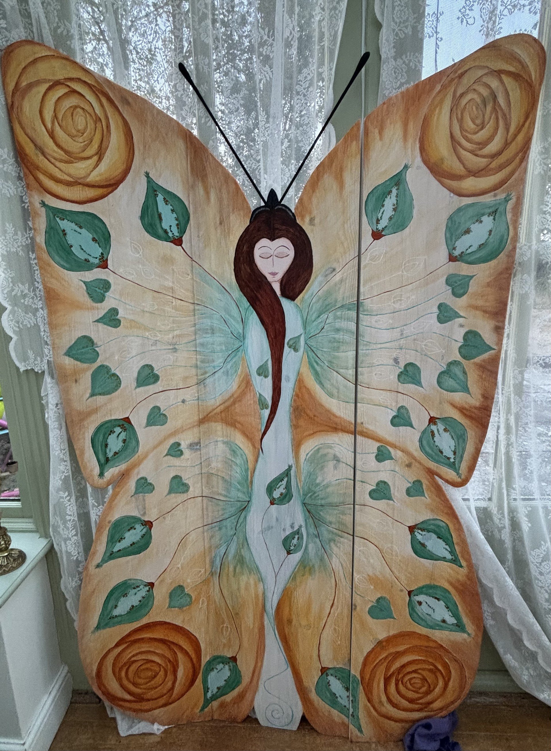 A large painted butterfly with a woman's face in the center, surrounded by green leaves and orange roses, with lace curtains in the background.