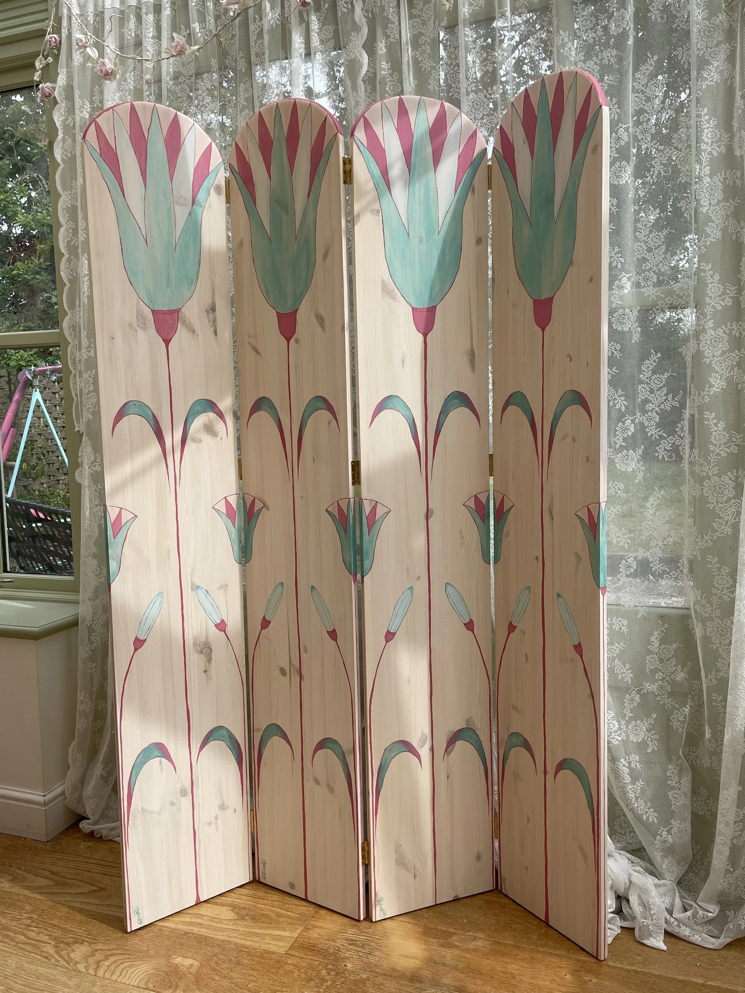 A decorative folding room divider with a painted floral design featuring large, stylized tulips in pink, turquoise, and green on a light wooden background, set indoors near lace curtains and a window.