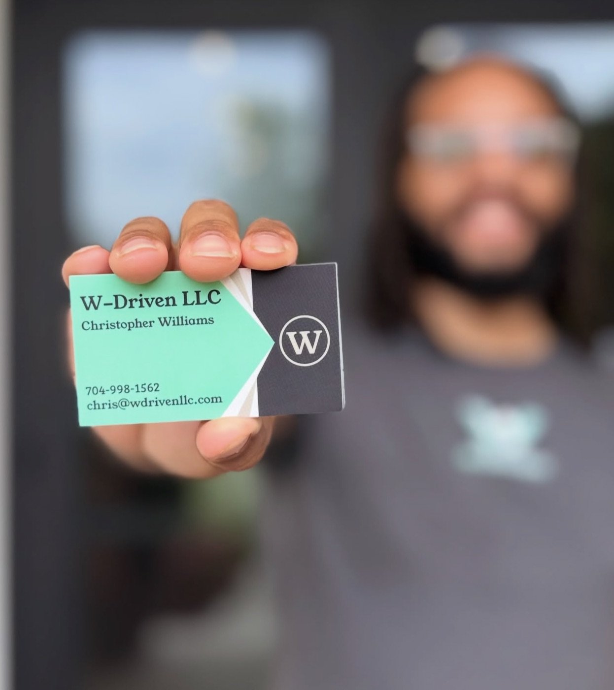 Person holding a business card for W-Driven LLC with contact information, blurred person in background.