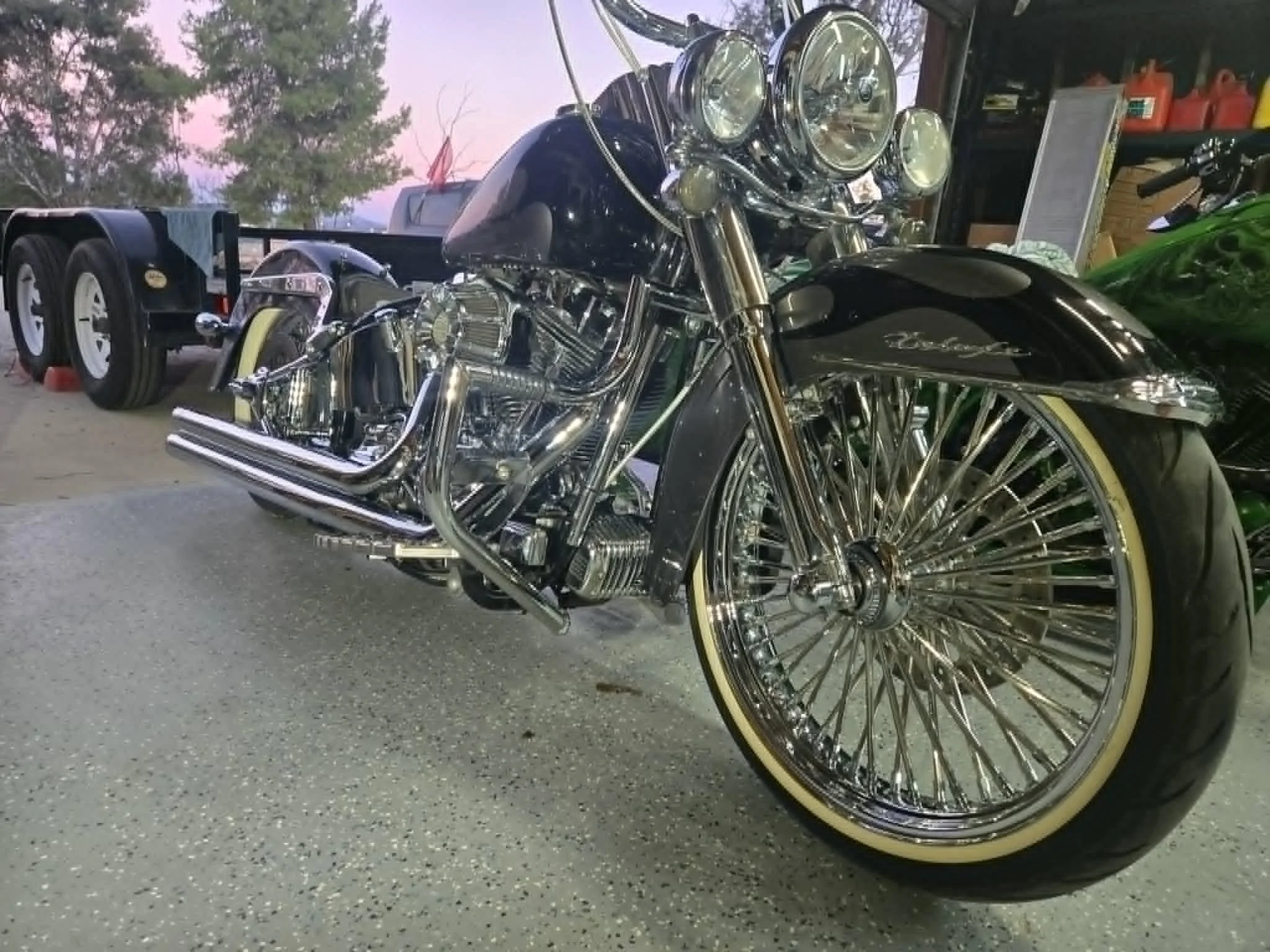 A black motorcycle with chrome details and cream-colored tires parked on a gravel surface, with a tree line and a pinkish sunset sky in the background.