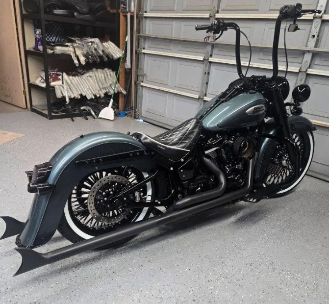 A custom black and teal motorcycle with long ape hanger handlebars, a quilted black seat, and black exhaust pipes parked in a garage.