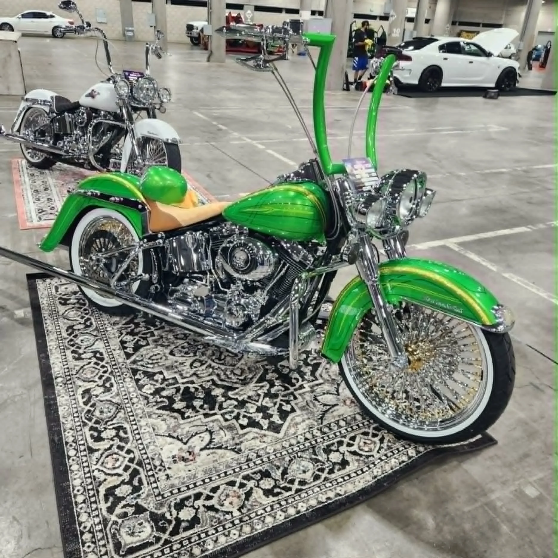 A shiny green motorcycle with chrome details displayed on a black and white patterned rug in a parking garage. Other vehicles and people are visible in the background.