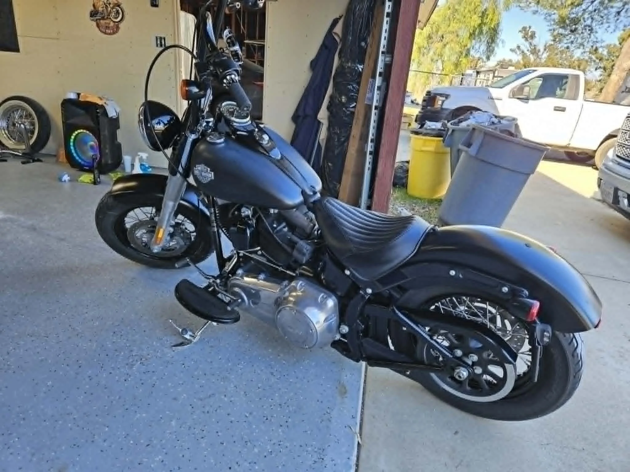 A black motorcycle parked in a garage, with various tools and items on shelves and the open garage door revealing a truck and trash bins outside.