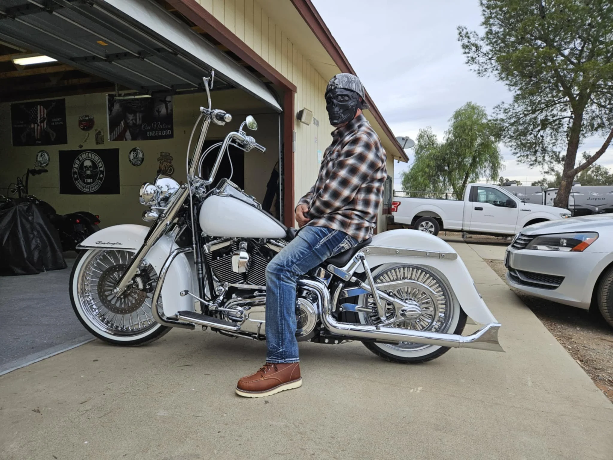 A person sitting on a white motorcycle in a driveway outside a house, wearing a plaid shirt, jeans, brown boots, a helmet, goggles, and a face mask.