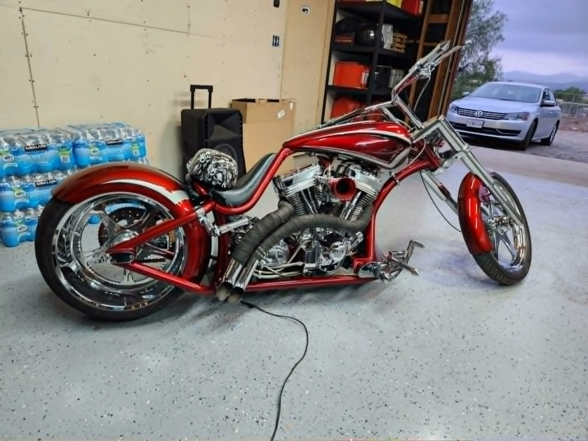 A red motorcycle with chrome accents parked in a garage, with bottled water stacked against the wall and shelving units in the background. A gray car is visible outside through the open garage door.