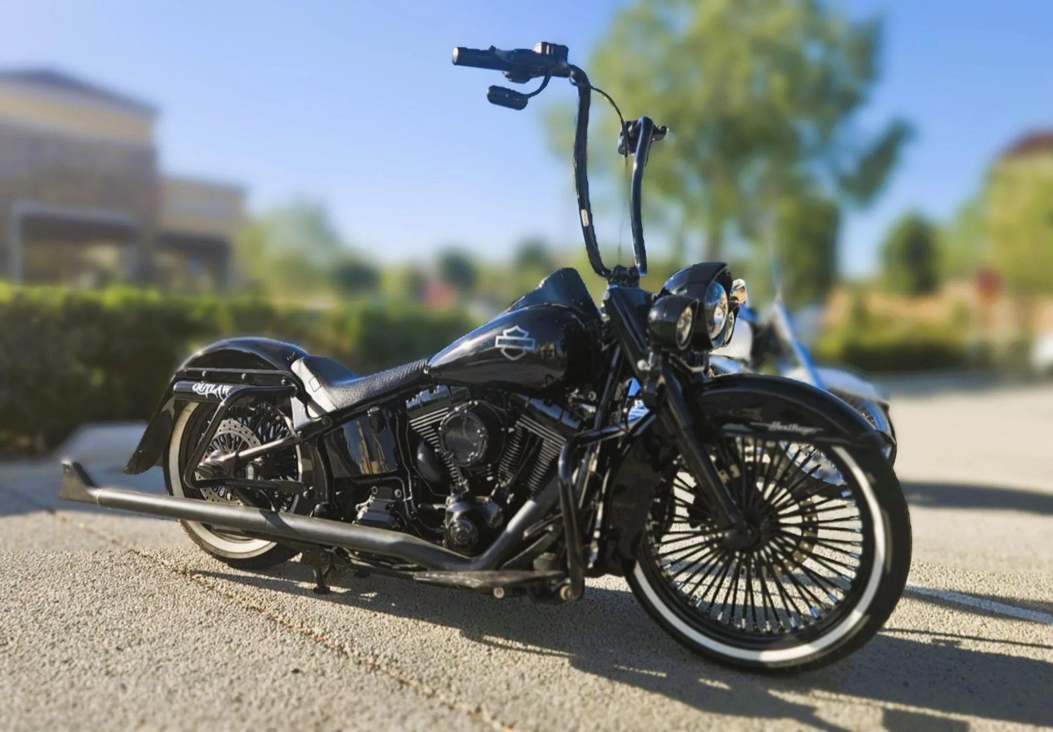 A black motorcycle with a custom handlebar designed for a chopper-style bike, parked on a paved surface outdoors with greenery and buildings in the background.
