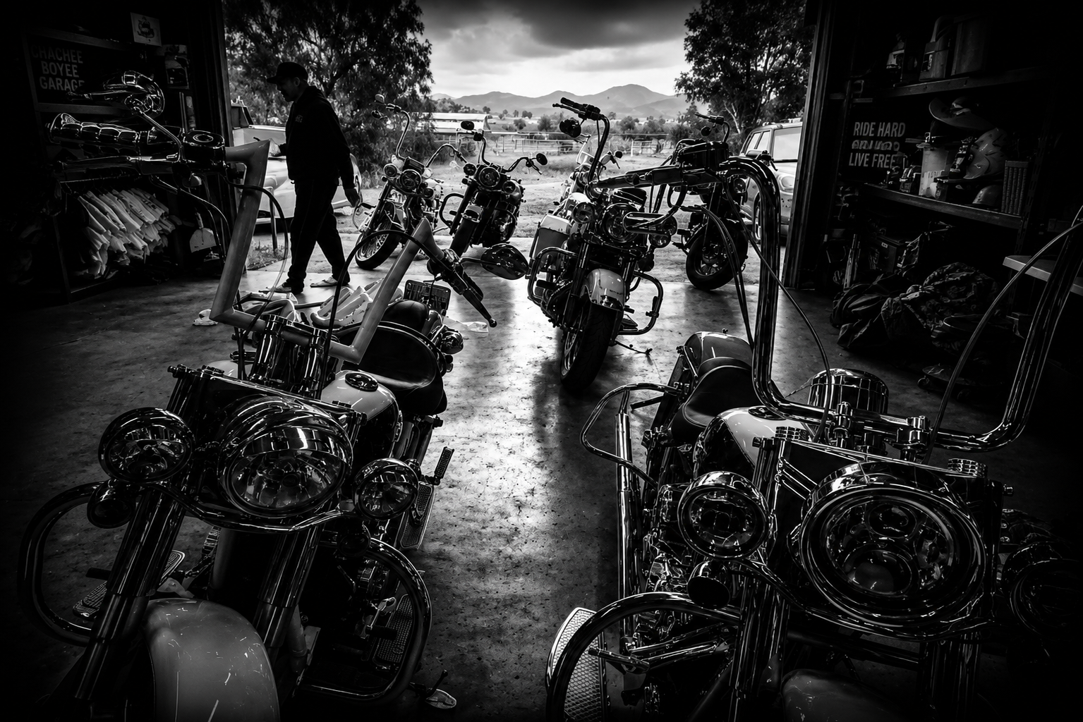 Black and white photo of a motorcycle garage with several parked motorcycles and a person inspecting one, open garage door revealing a rural landscape with mountains and trees in the background.