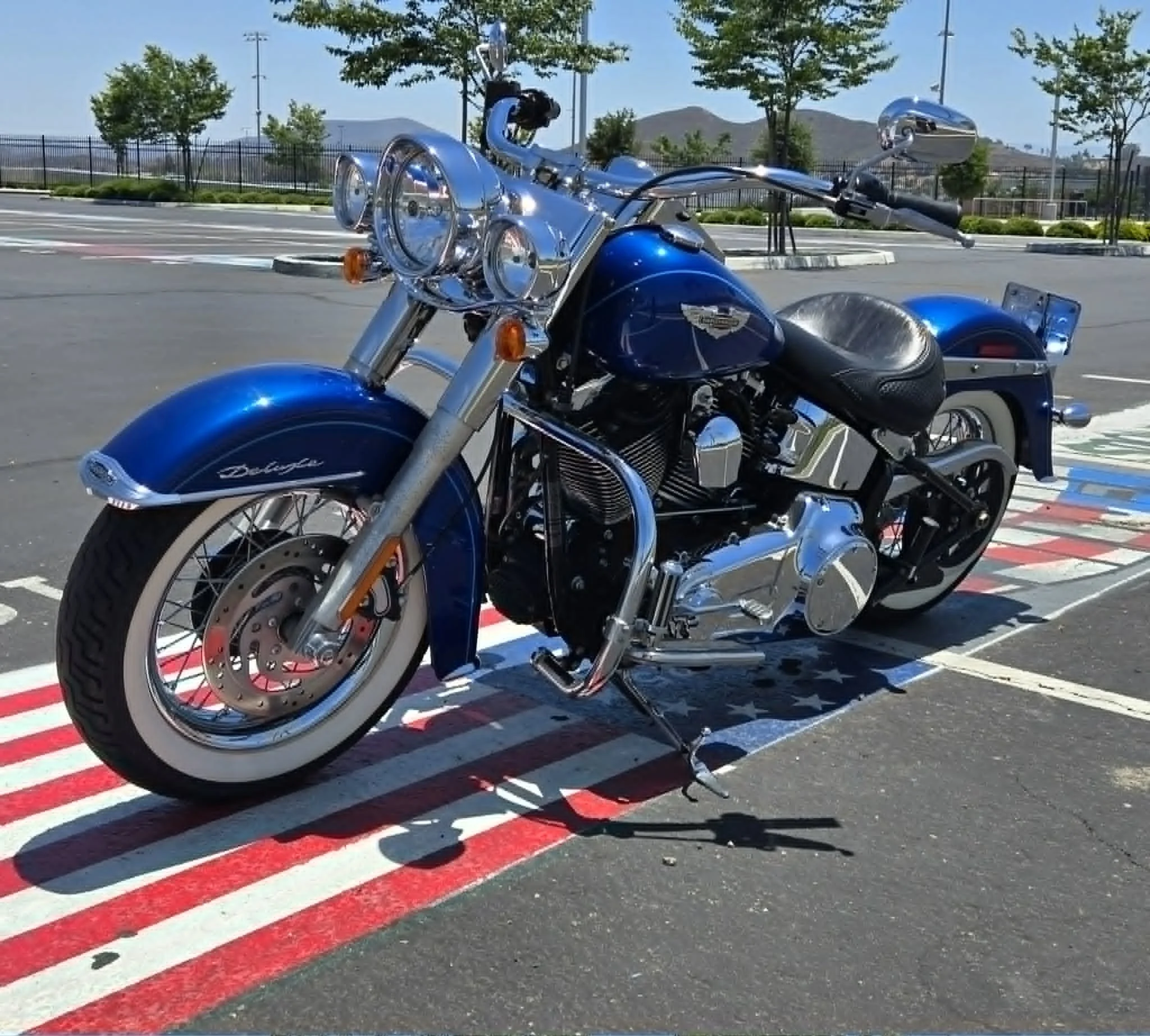 A blue motorcycle parked in a parking lot, with painted red and white lines on the asphalt and a fence in the background.