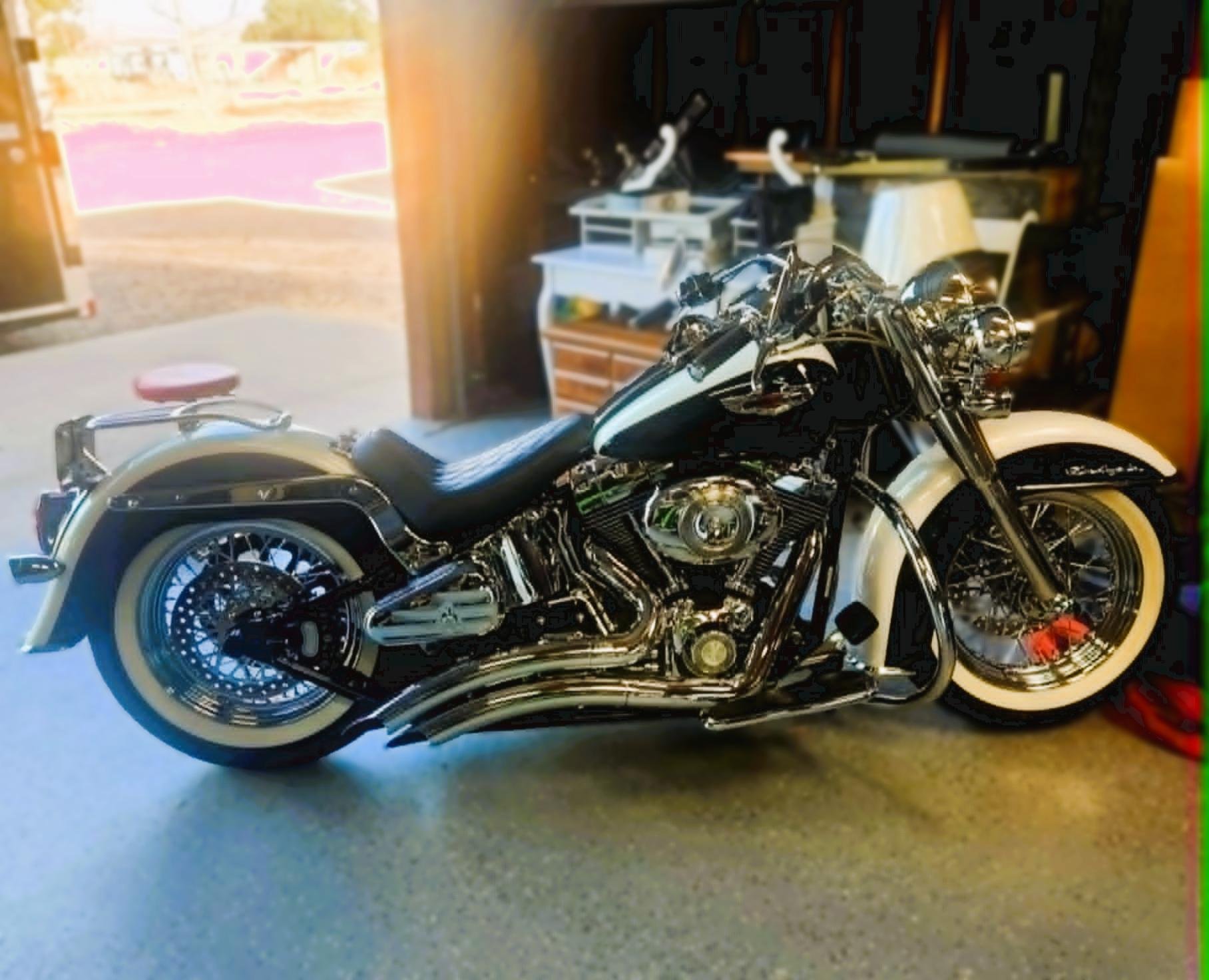 A Harley-Davidson motorcycle with a white-walled tire, parked inside a garage. The garage has a workbench with various tools and equipment, with sunlight coming in through an open door.