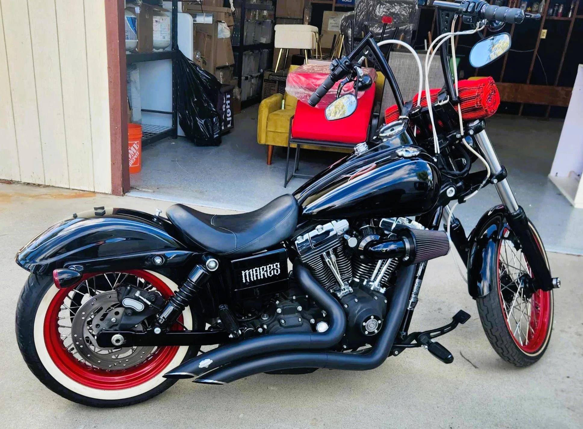A black motorcycle with red rims parked outside a store. The store's interior shows various furniture and boxed items, with a yellow chair, a red rolled-up mat, and other miscellaneous items visible.