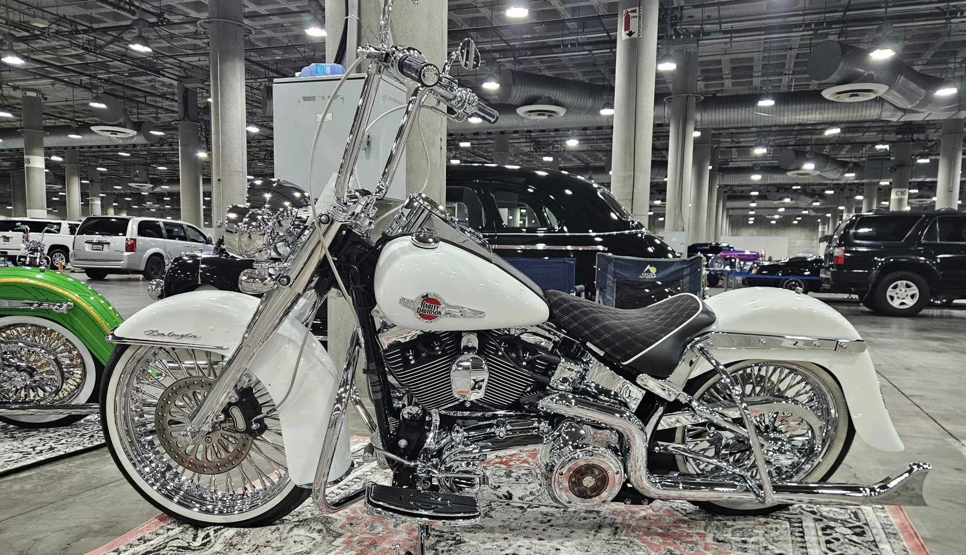 A custom white Harley Davidson motorcycle with chrome accents and a black and white diamond patterned seat displayed in an indoor parking garage with other vehicles in the background.