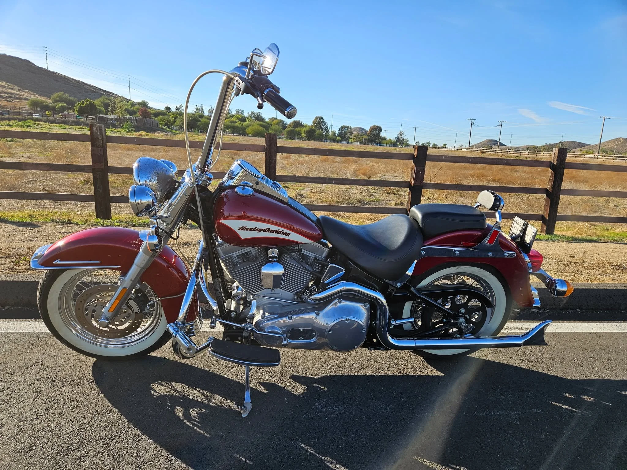 Red Harley Davidson motorcycle parked on a paved road with a wooden fence and a landscape of hills, trees, and utility poles under a clear blue sky.