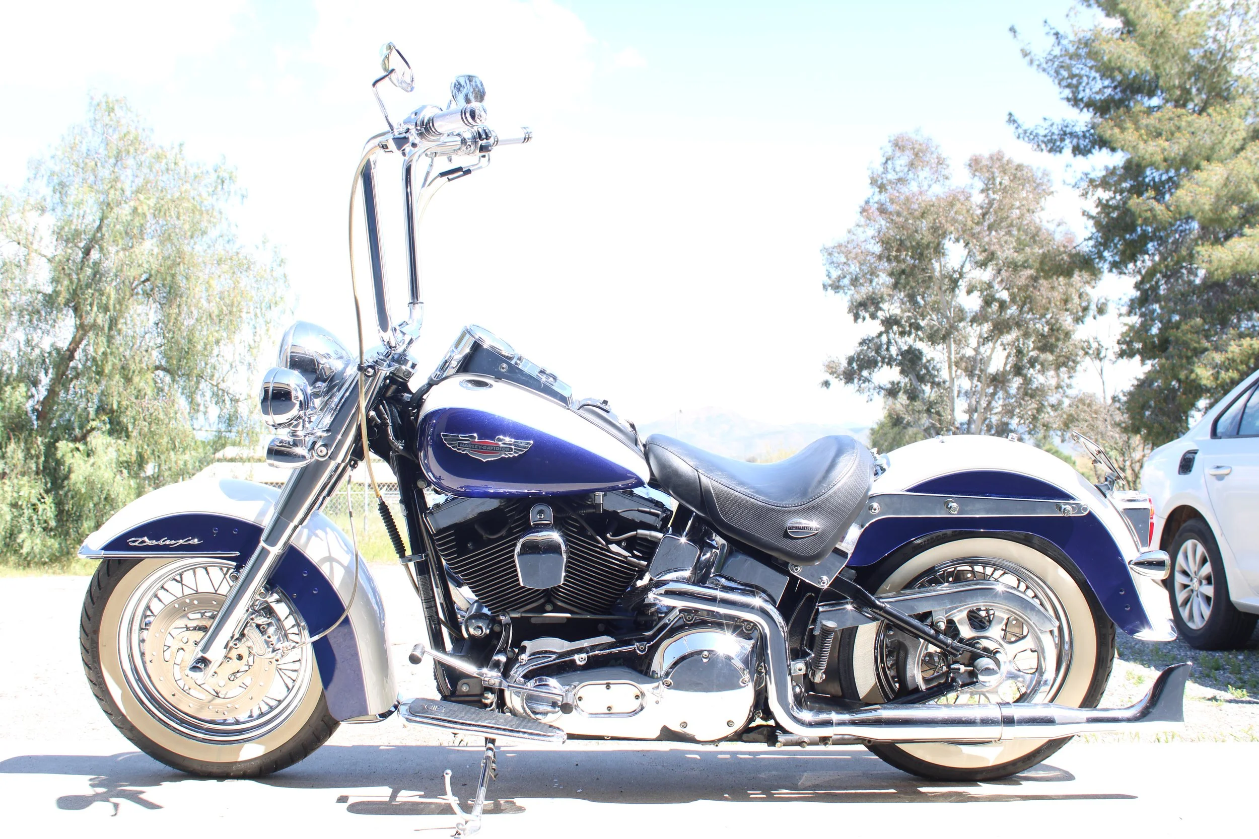 A blue and chrome Harley-Davidson motorcycle parked outdoors on a sunny day with trees and a white vehicle in the background.