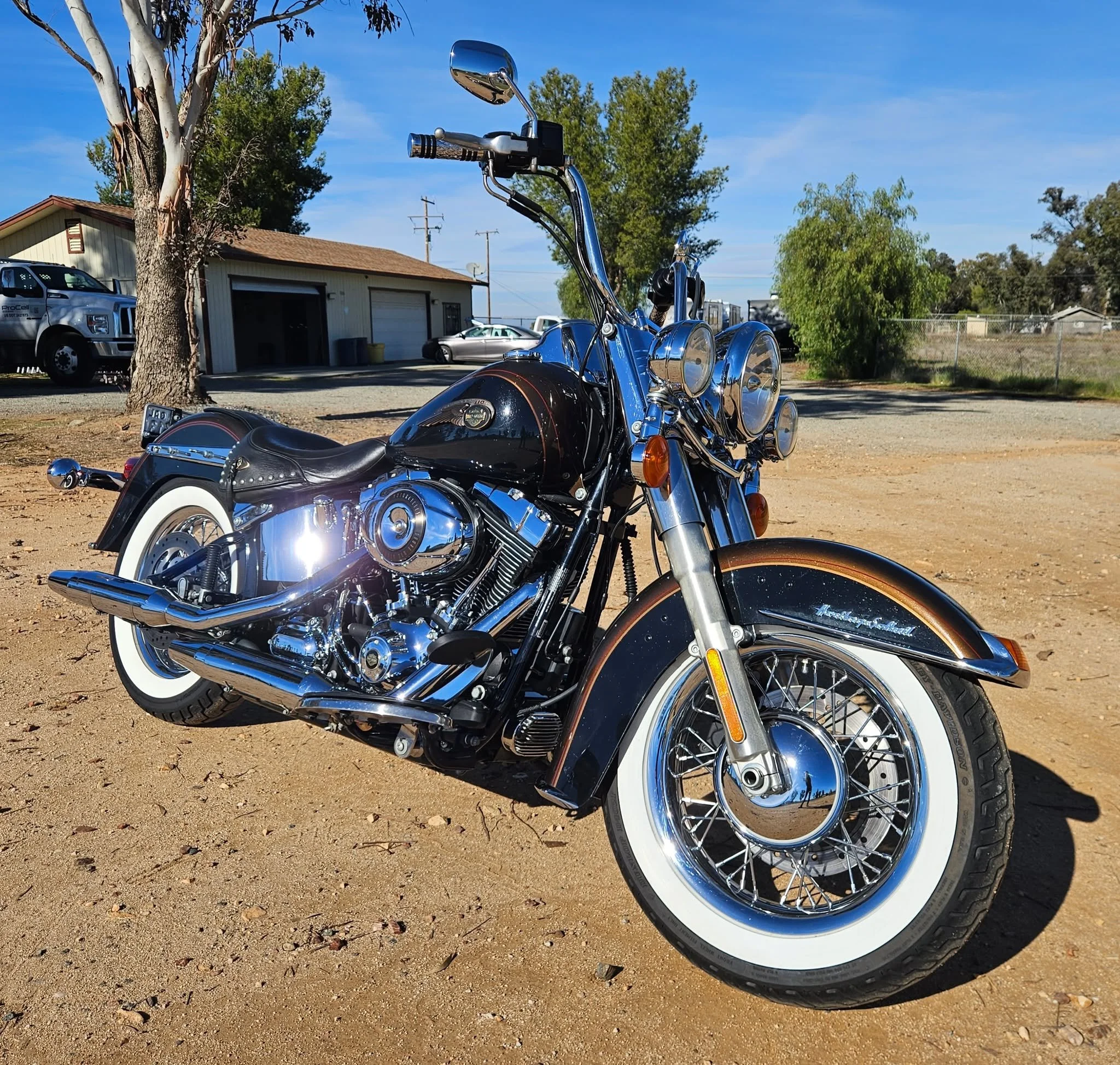 A black and chrome Harley-Davidson motorcycle with white-wall tires parked on a dirt surface, with trees, a building, and parked vehicles in the background under a clear blue sky.