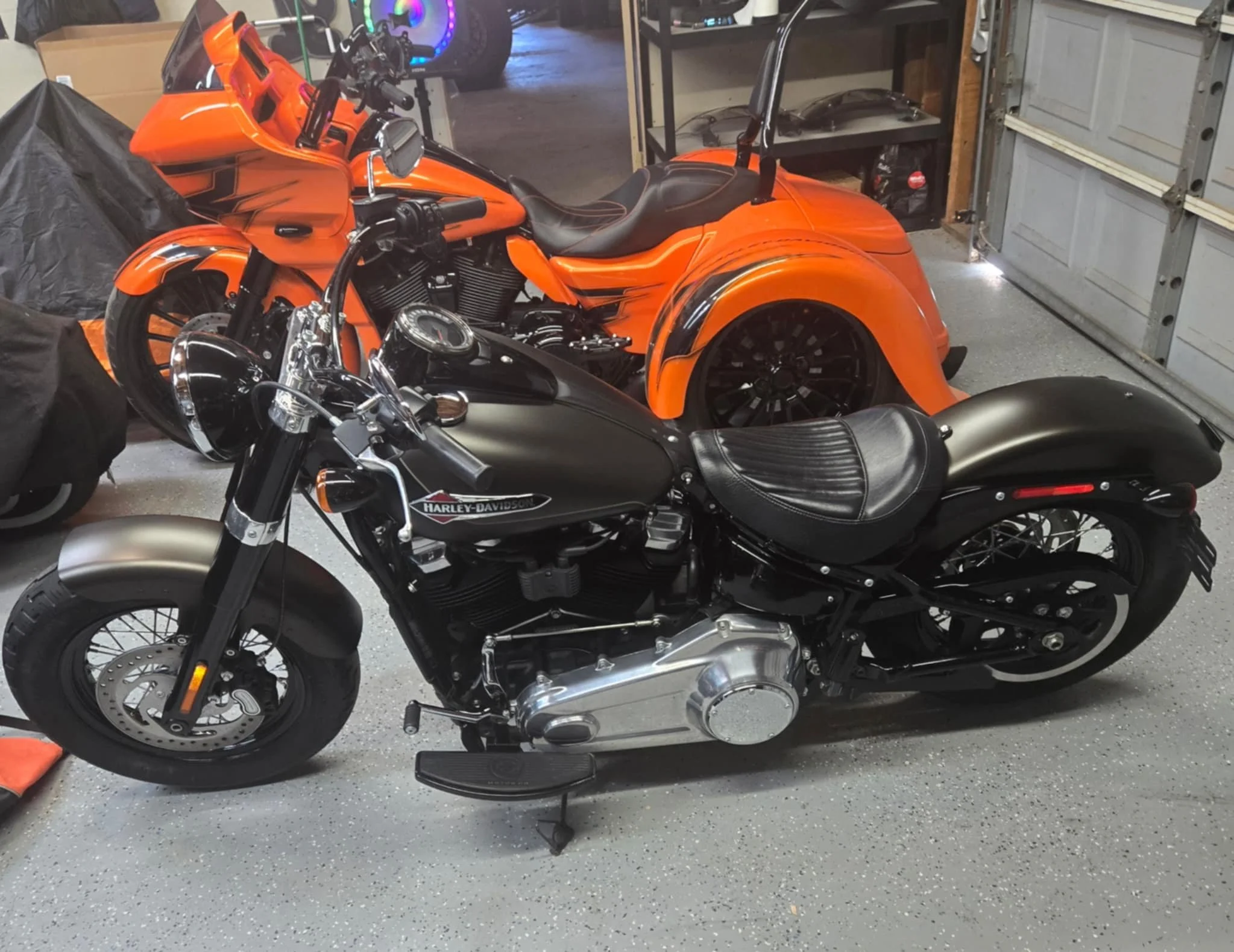A garage interior with two motorcycles, an orange three-wheeled motorcycle and a black Harley-Davidson motorcycle, parked on a speckled gray floor near a gray garage door and black shelving units.