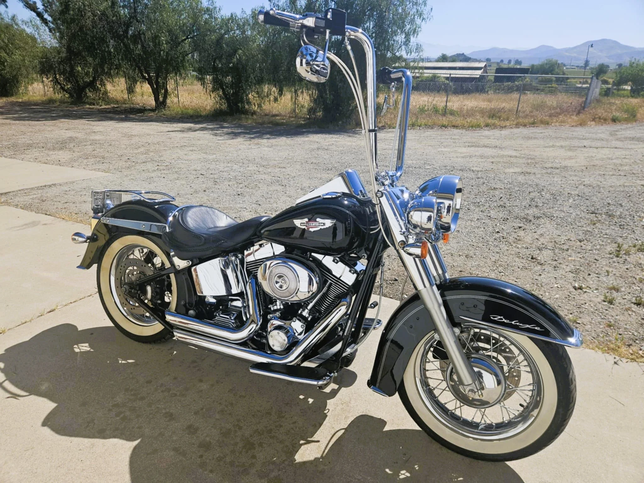 A black and chrome Harley-Davidson motorcycle parked on a concrete sidewalk with a dirt lot and trees in the background.