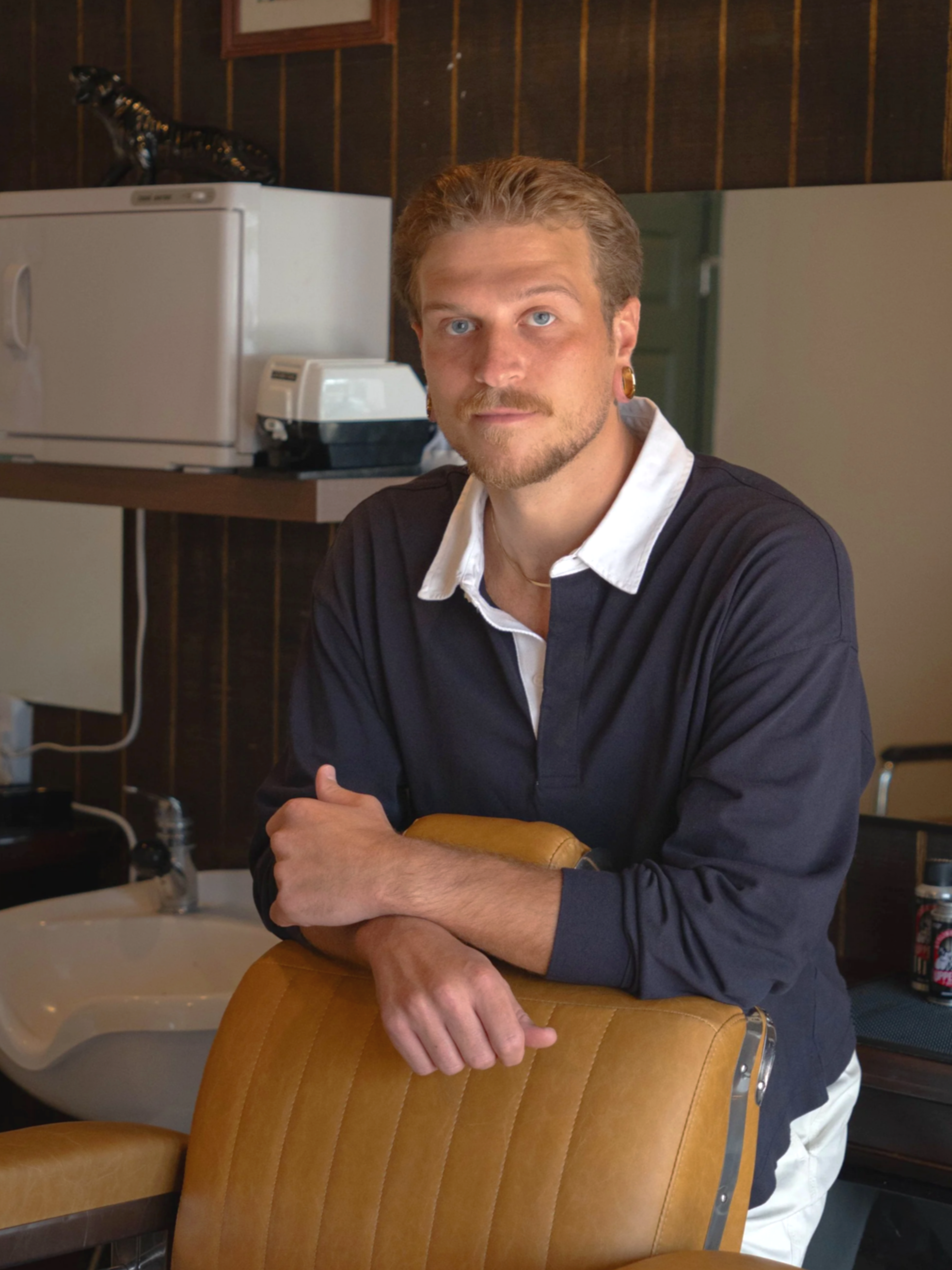 barber standing behind his chair, hot towel cabinet and lather machine can be seen behind him