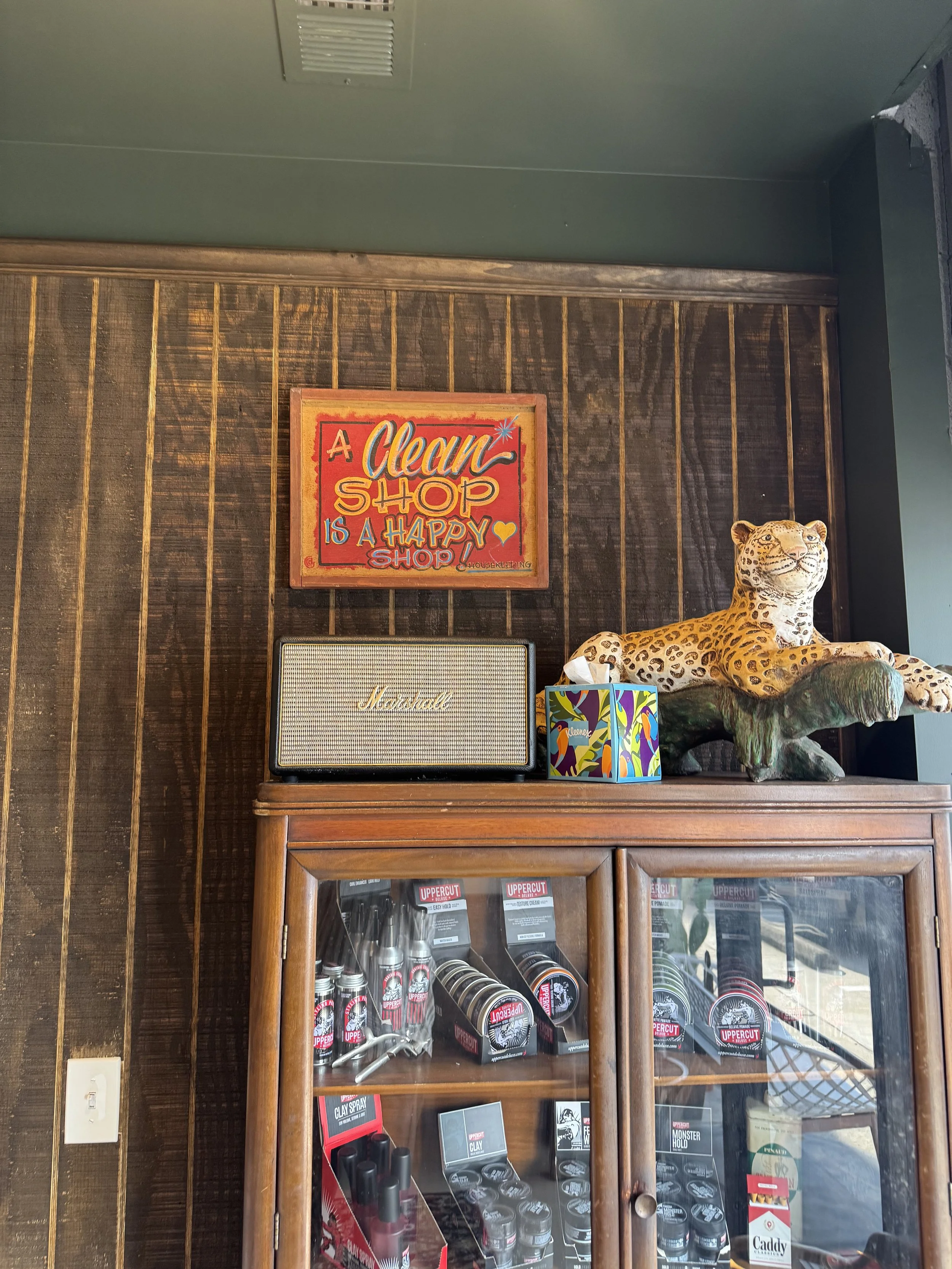 A wooden display cabinet filled with various small spray paint cans and related products. On top of the cabinet, there's a decorative leopard sculpture on the right and a colorful tissue box. Mounted on the wall above it is a sign that says, "A Clean Shop is a Happy Shop!" with vibrant colors.