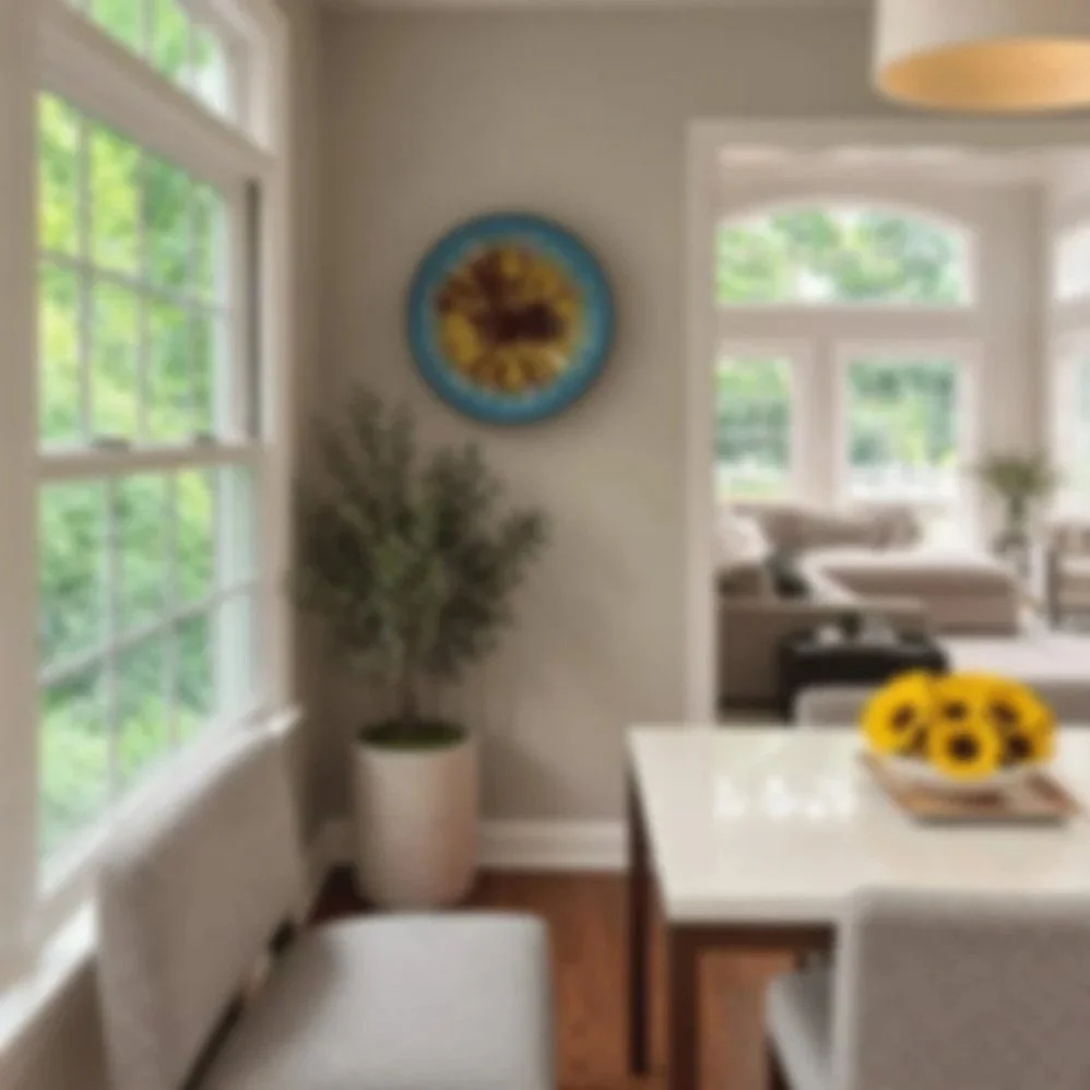 Interior of a bright dining room with large windows, a white dining table with a sunflower centerpiece, a bench, a potted plant, and a wall clock, leading to a living room area with sofas and large windows.