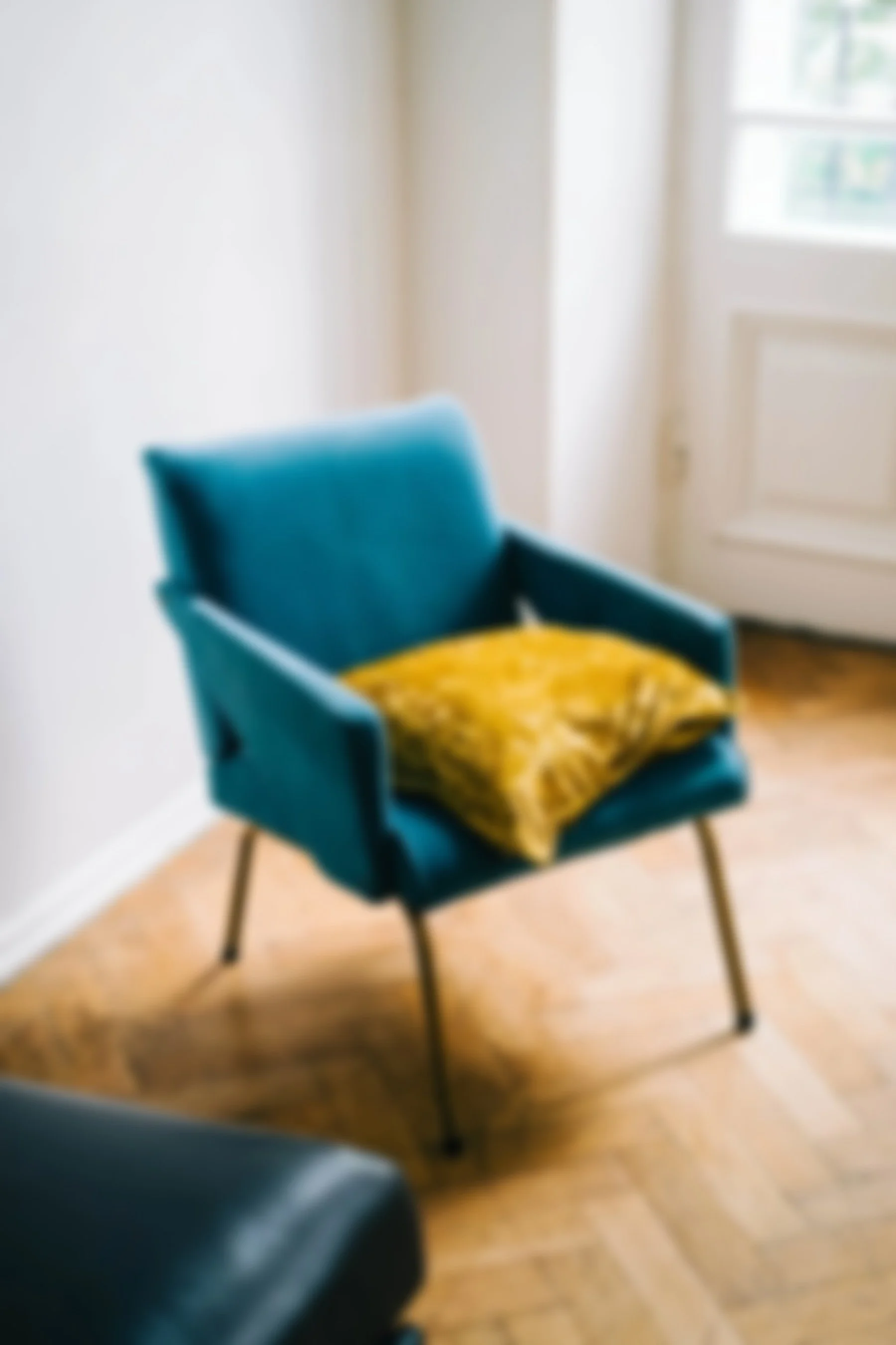 A blue armchair with wooden legs and a yellow patterned throw pillow on it, situated on a wooden floor near a white wall and window.