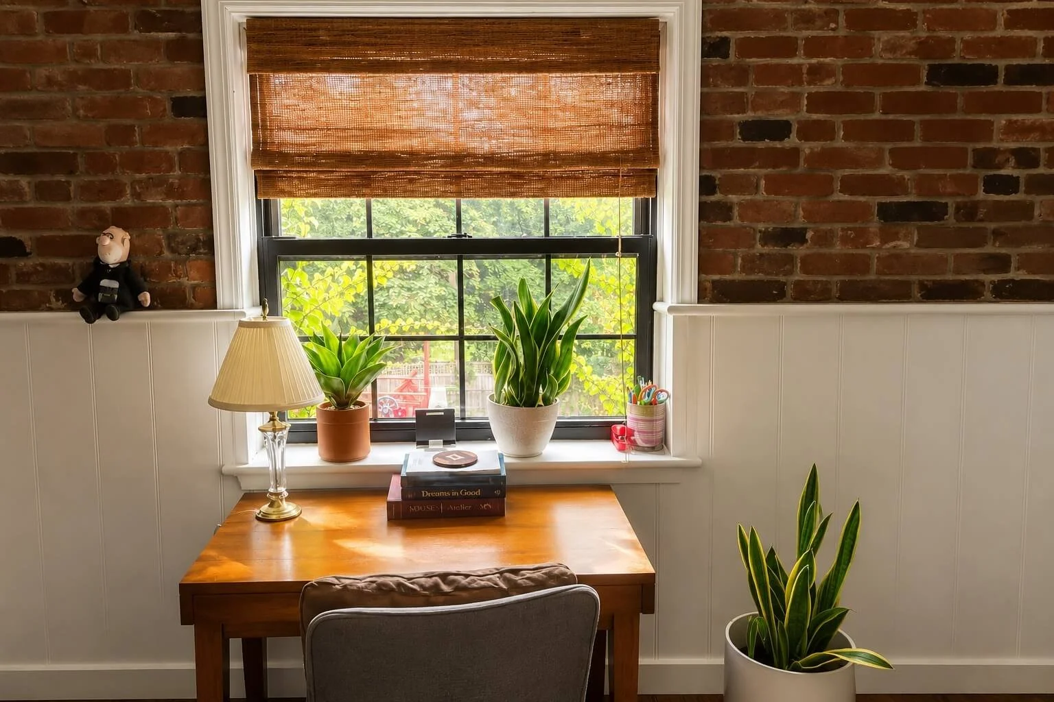 A cozy home office corner with a wooden desk, a gray chair, and a window with a bamboo blind. The desk has a table lamp, two potted plants, a stack of books, and a small container with pens and scissors. The background features a brick wall and white wainscoting.