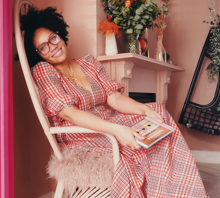 A woman with curly hair and glasses sitting in a cozy room, smiling while holding a book. She is wearing a red checkered dress and sitting in a light pink chair with a fluffy cushion. There is a fireplace with colorful flowers on the mantel and a tilted black chair in the background.
