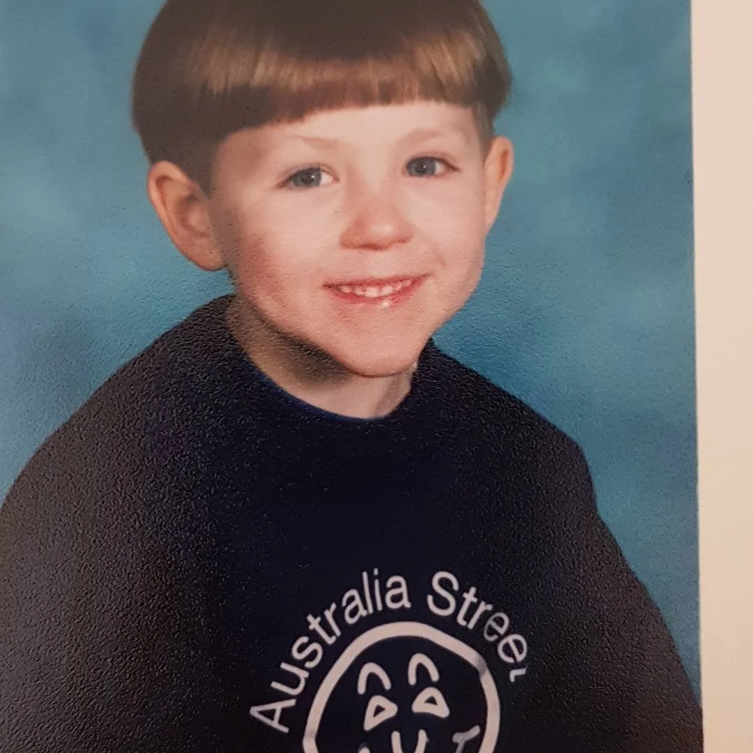 Portrait of a young boy with short brown hair, blue eyes, and a big smile, wearing a black sweatshirt with a white logo in front that says "Australia Street."