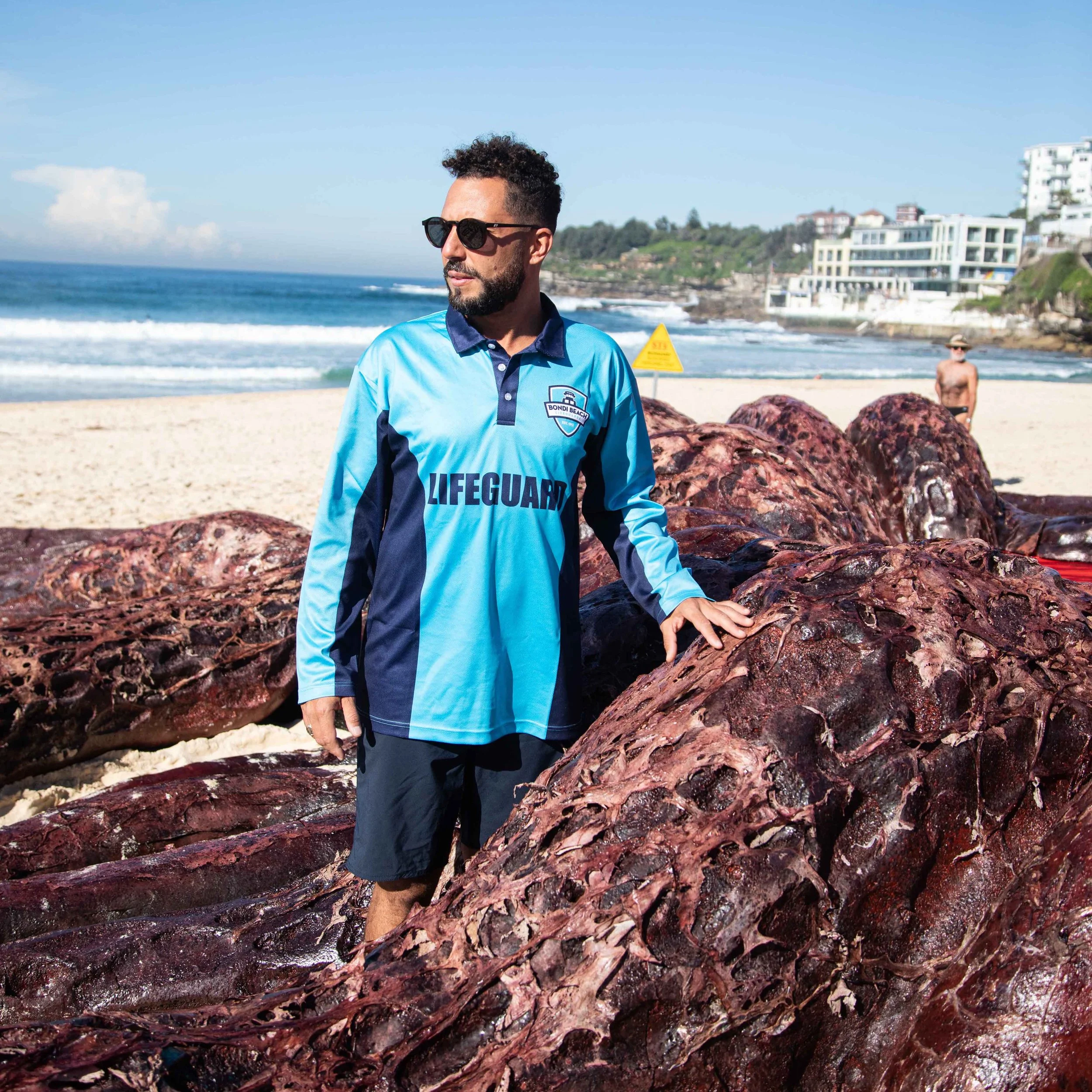 A man wearing sunglasses and a Lifeguard shirt standing on a beach with large red rocks, ocean waves, and buildings in the background.