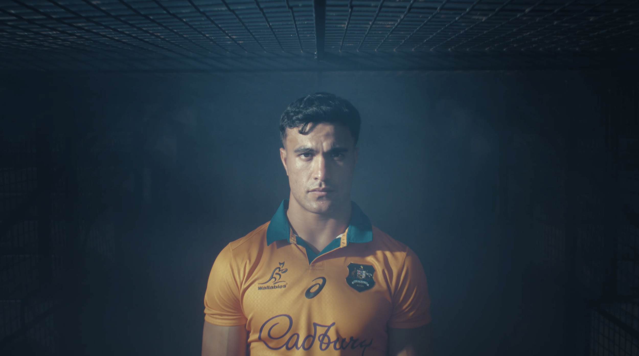 A male athlete in a yellow sports jersey with sponsor logos, standing in a dark tunnel with a grid ceiling, looking serious.
