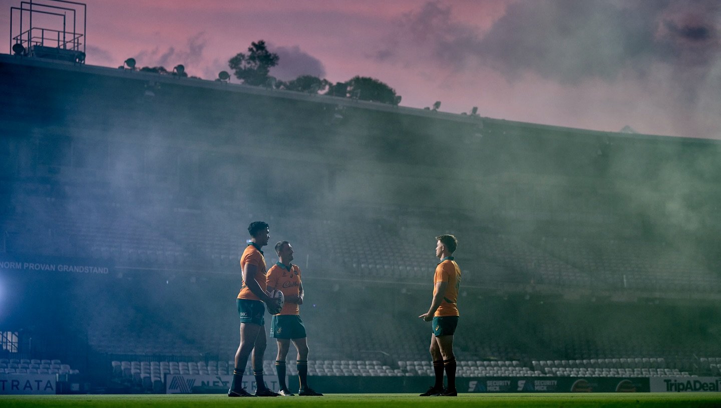 Three rugby players in orange jerseys and black shorts standing on a field, facing each other, with a stadium and cloudy sky in the background.
