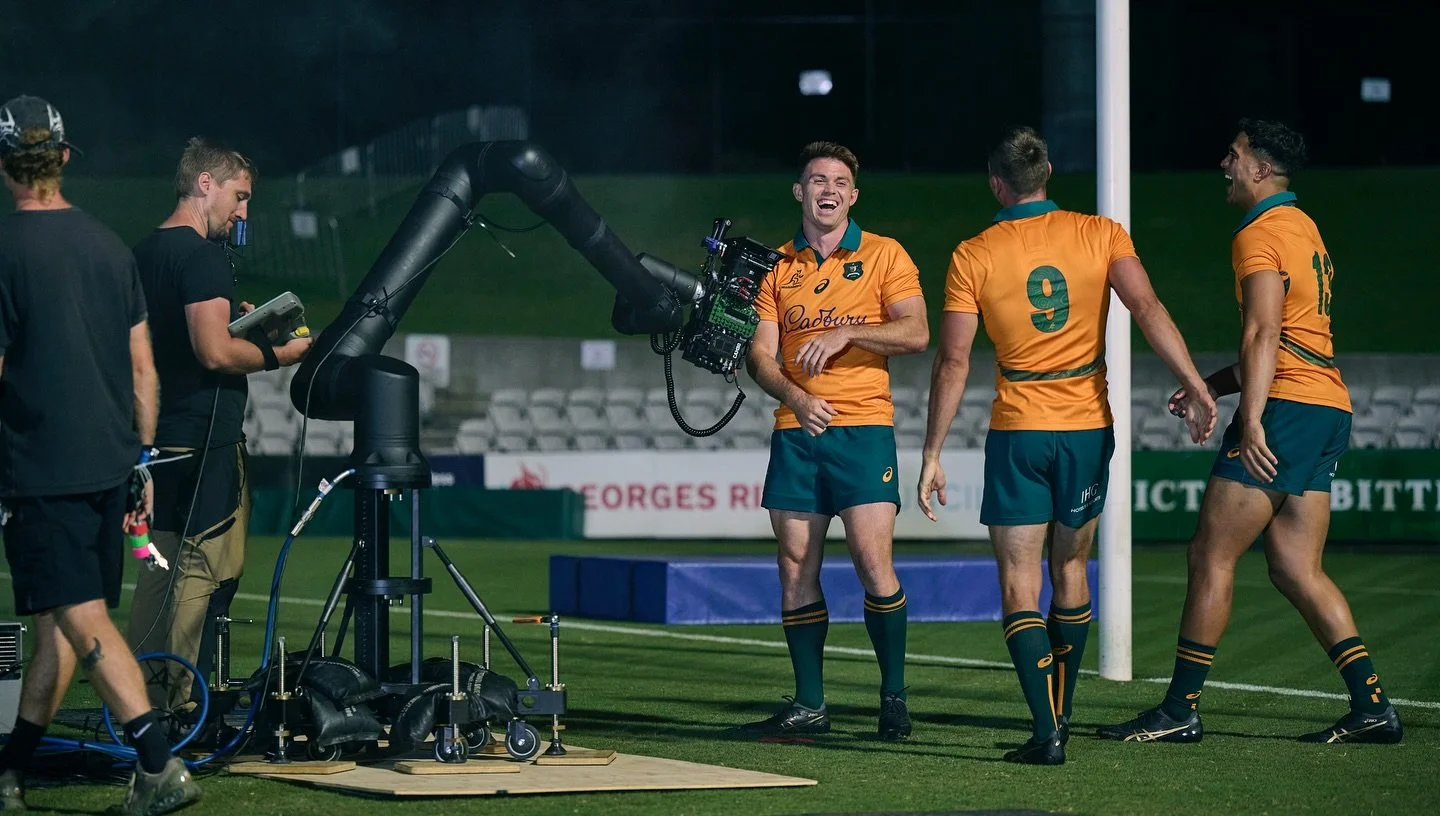 Australian rugby players being filmed during a match at night. Three players in orange and green uniforms are near a goalpost, two are seen from behind and one from the front, smiling and laughing. Two crew members with filming equipment are on the left side of the image.