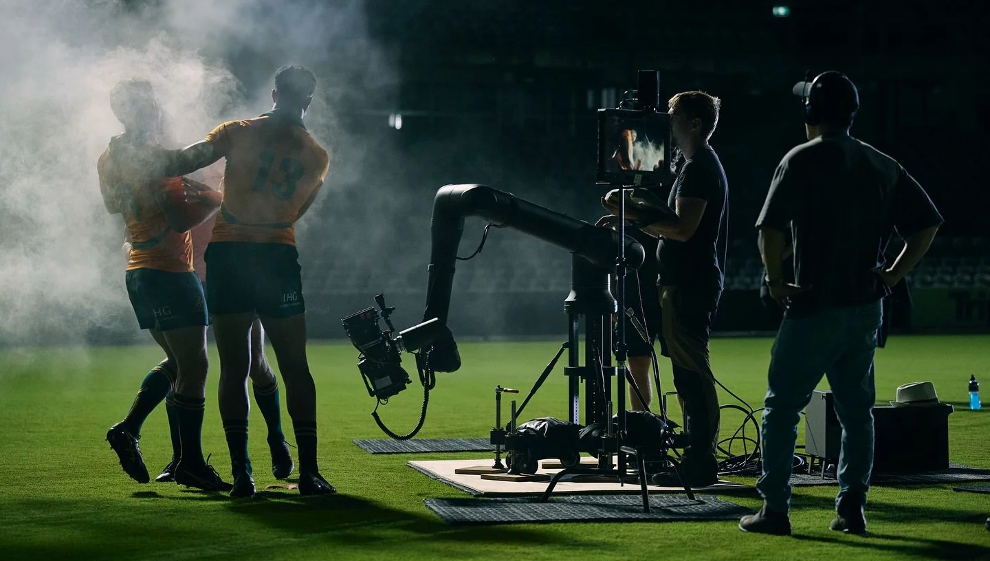Two athletes wearing sports uniforms are on a sports field, with one offering something to the other, while a production crew films them with a camera on a rig, under artificial lighting in a dark setting.
