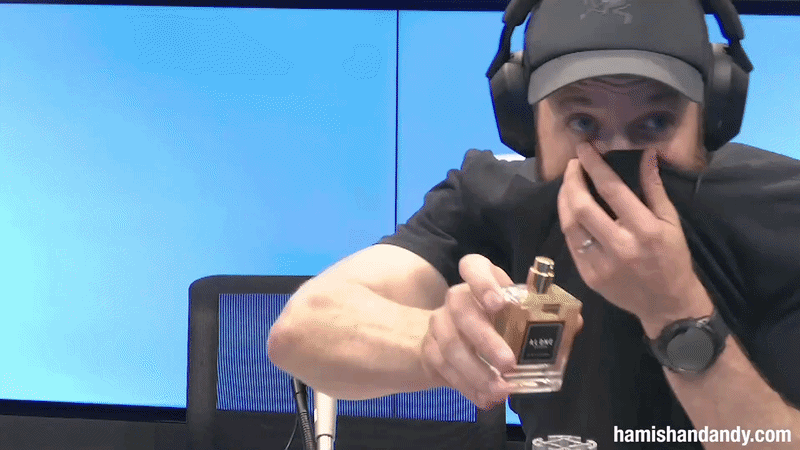 A man wearing headphones and a cap covering his nose with one hand while holding a perfume bottle pointed toward the camera in a recording studio.