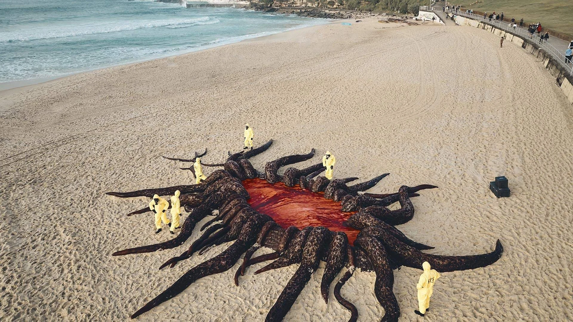 A large sea monster sculpture on a sandy beach, with multiple humans in yellow hazmat suits surrounding it, some examining and some taking photos. The sculpture has a long, dark, textured body with numerous tentacle-like appendages, and a red, bloody interior exposed.