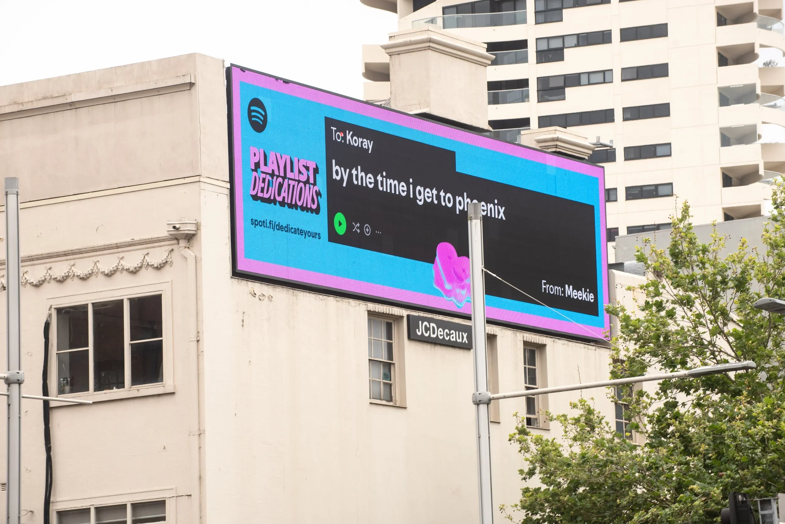Large digital billboard on a building displaying a Spotify playlist dedication message with a bright blue and pink theme, trees partially visible in the foreground.