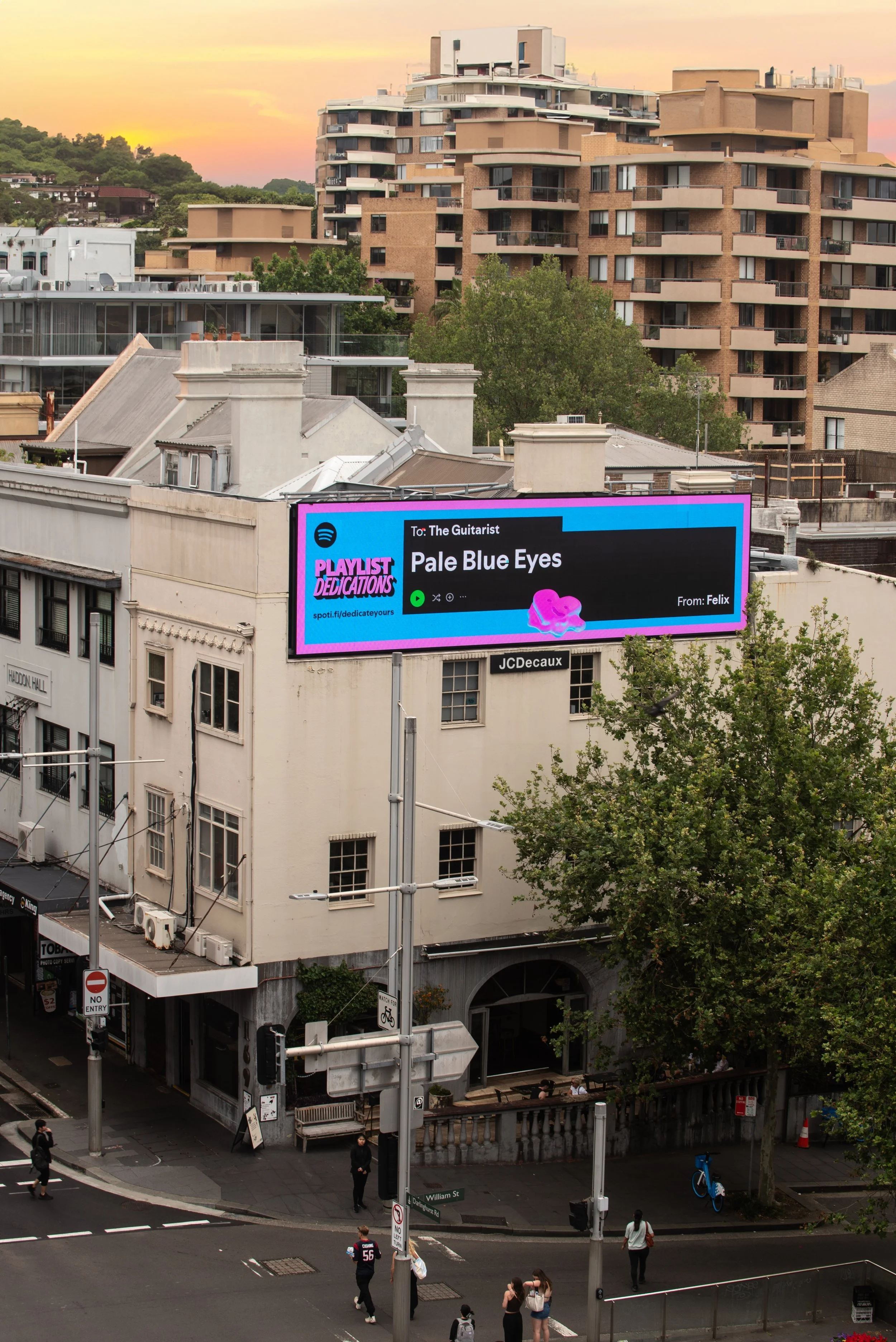 A city street scene at sunset with a billboard and several pedestrians. The billboard displays a music playlist titled "Pale Blue Eyes" sent to 'The Guitarist' from 'Felix,' with a pink abstract design.