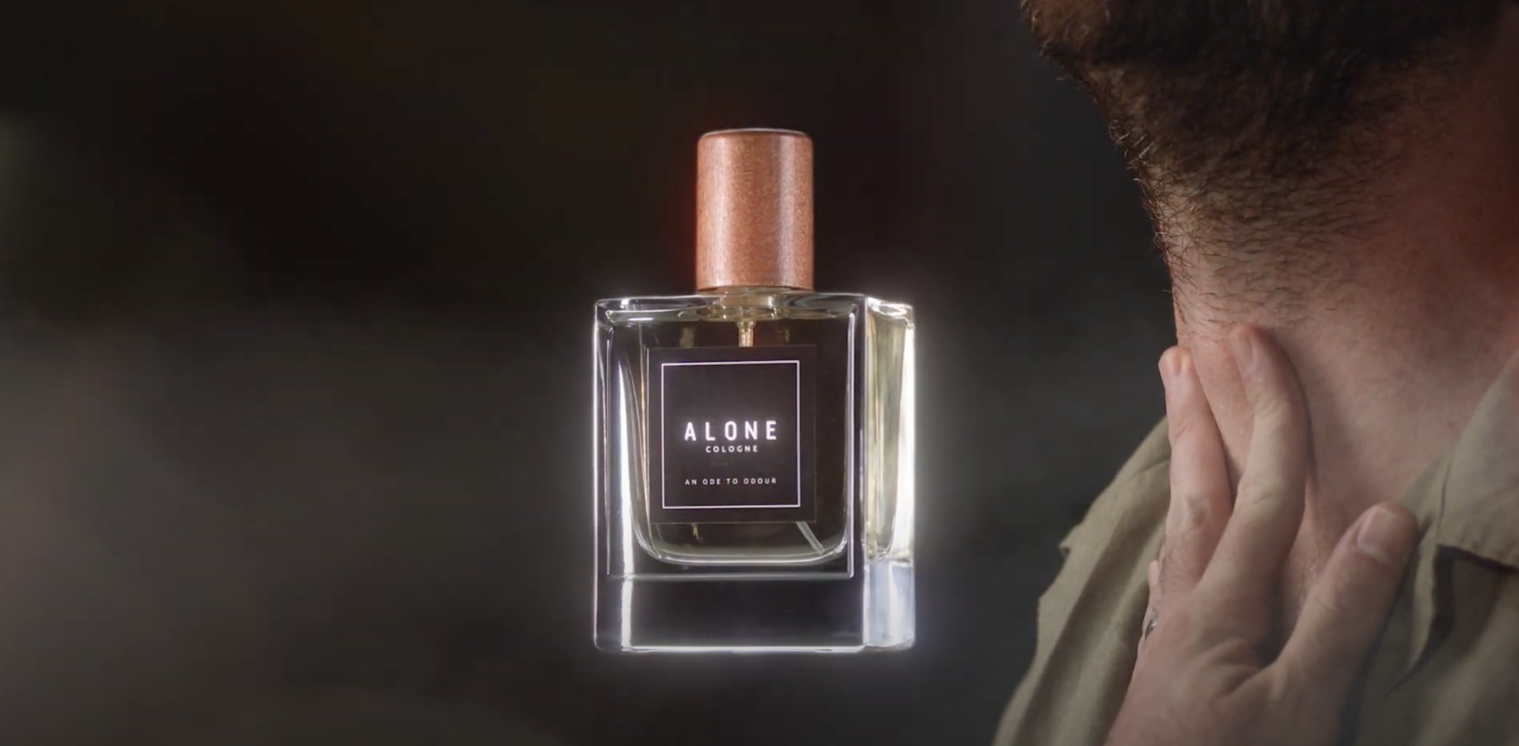 A man smells a bottle of cologne labeled 'Alone Cologne' with the subtitle 'An Ode to Odour' on a reflective surface.