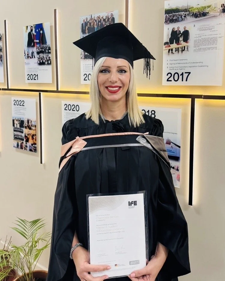 Woman in graduation cap and gown holding diploma, standing in front of a wall with photo posters from different years.