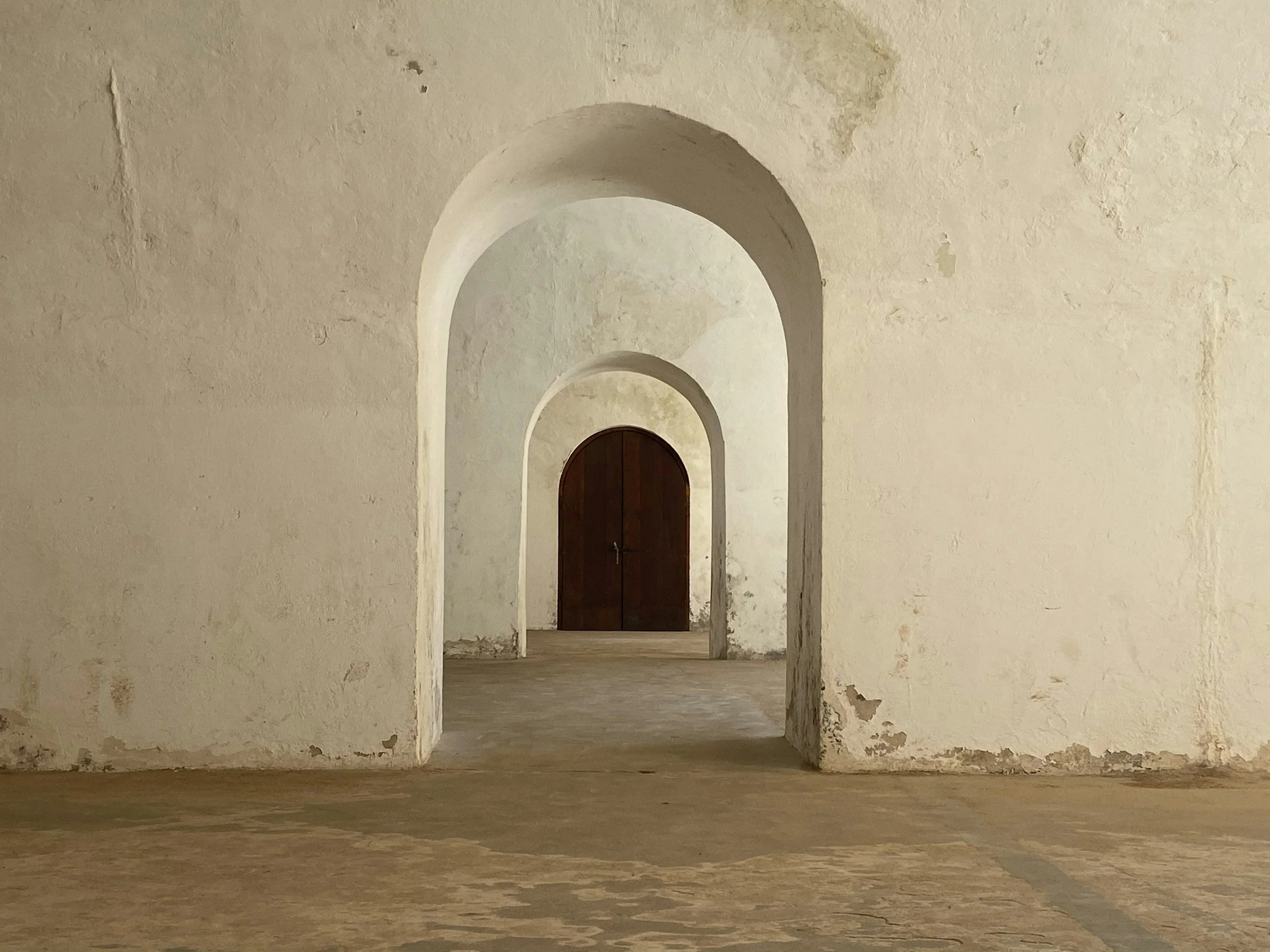 A series of arched doorways in an old, weathered stone wall with a wooden door at the far end.