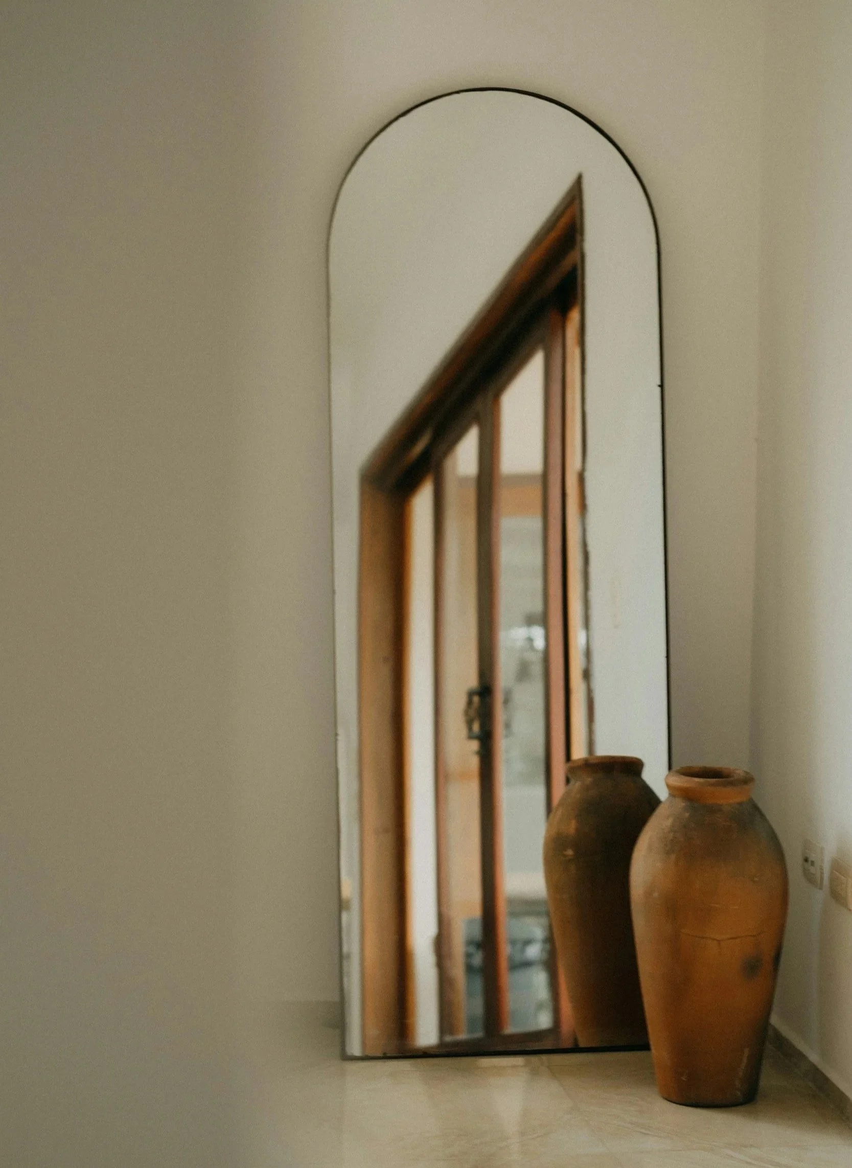 A tall mirror leaning against a wall, with a wooden frame, reflecting part of a staircase. Two large brown clay pots are placed on the floor next to the mirror.